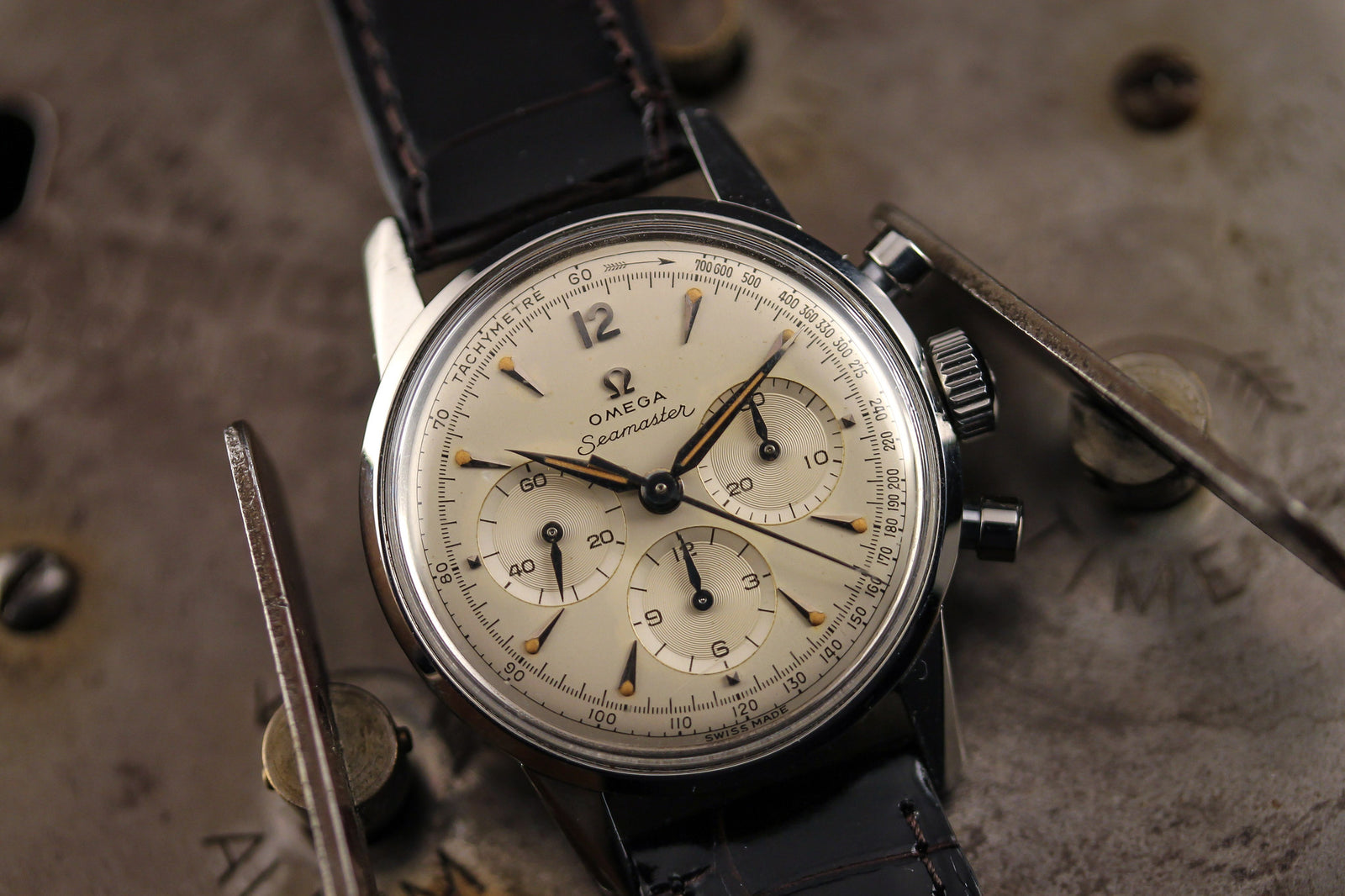 An Omega Seamaster Chronograph Calibre .321 - 1960 with a cream dial, black leather strap, and three subdials lies on an industrial metallic surface accented by screws and levers.