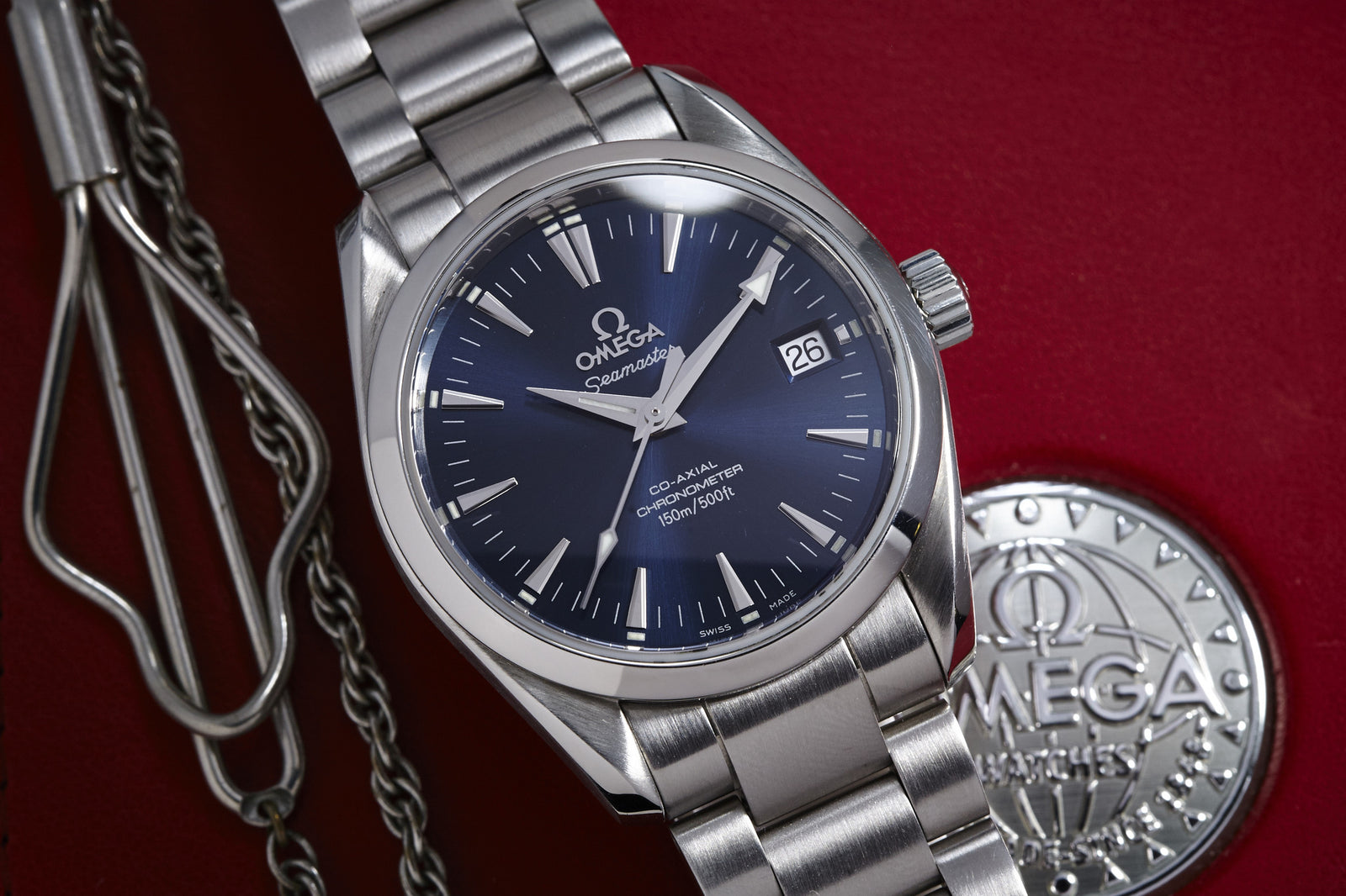 An Omega Seamaster Co-Axial Ref. 2504 Box & Papers wristwatch with a 36mm stainless steel case, blue dial, and date window is shown on a red surface beside an Omega medallion and a metal clasp with chain.