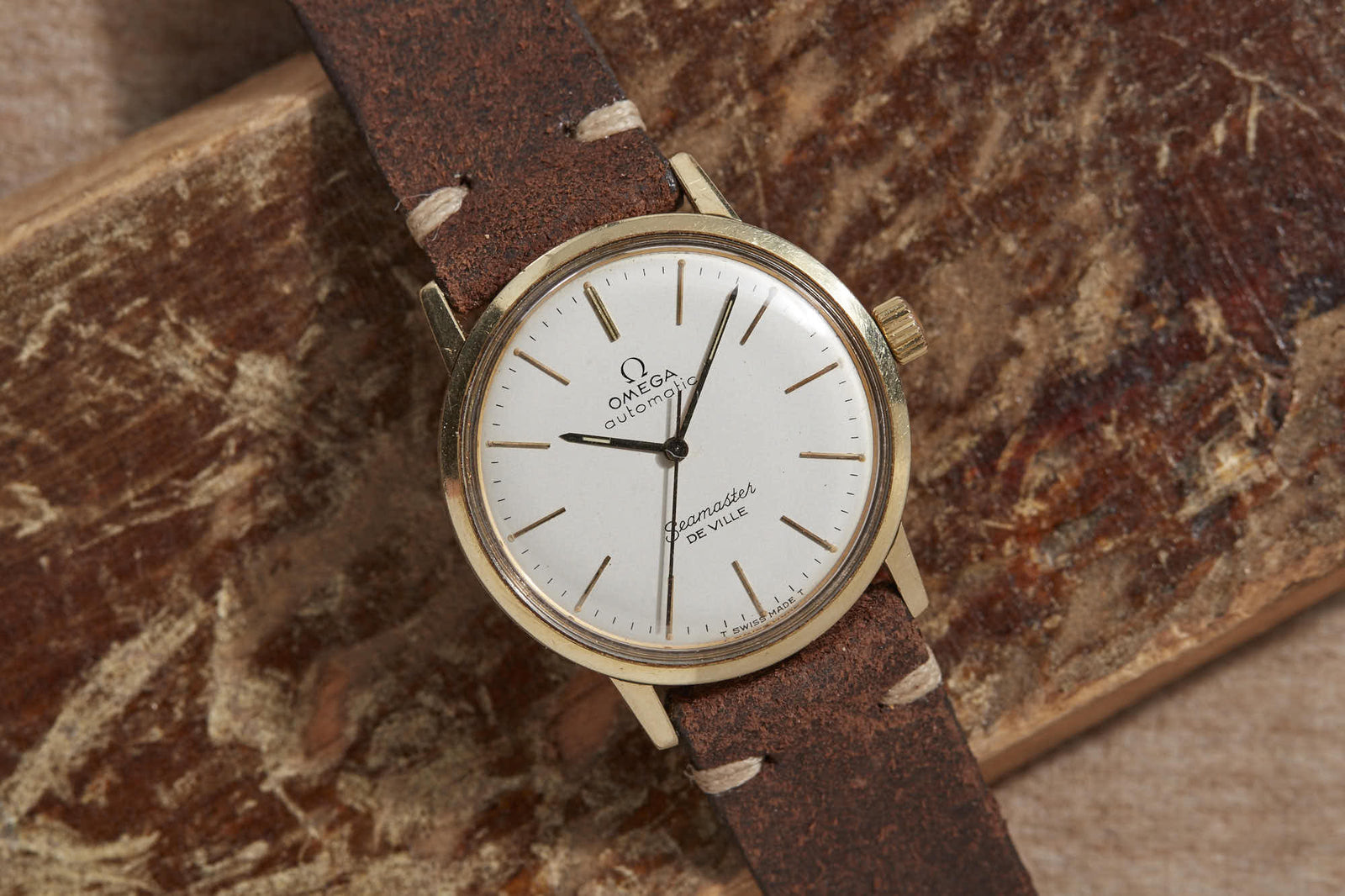 An Omega Seamaster De Ville wristwatch by Omega, featuring a white dial, gold case, and brown leather strap, displayed on a textured wooden surface—a classic statement of timeless luxury.