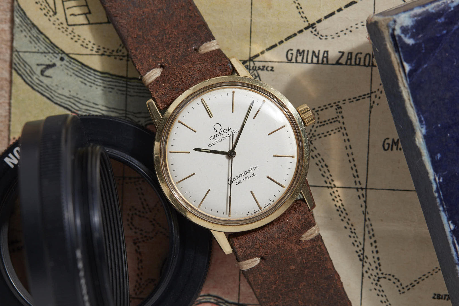 An Omega Seamaster De Ville by Omega with a brown leather strap rests on an old map beside a camera lens and blue book, showcasing its white dial with gold hour markers and hands.