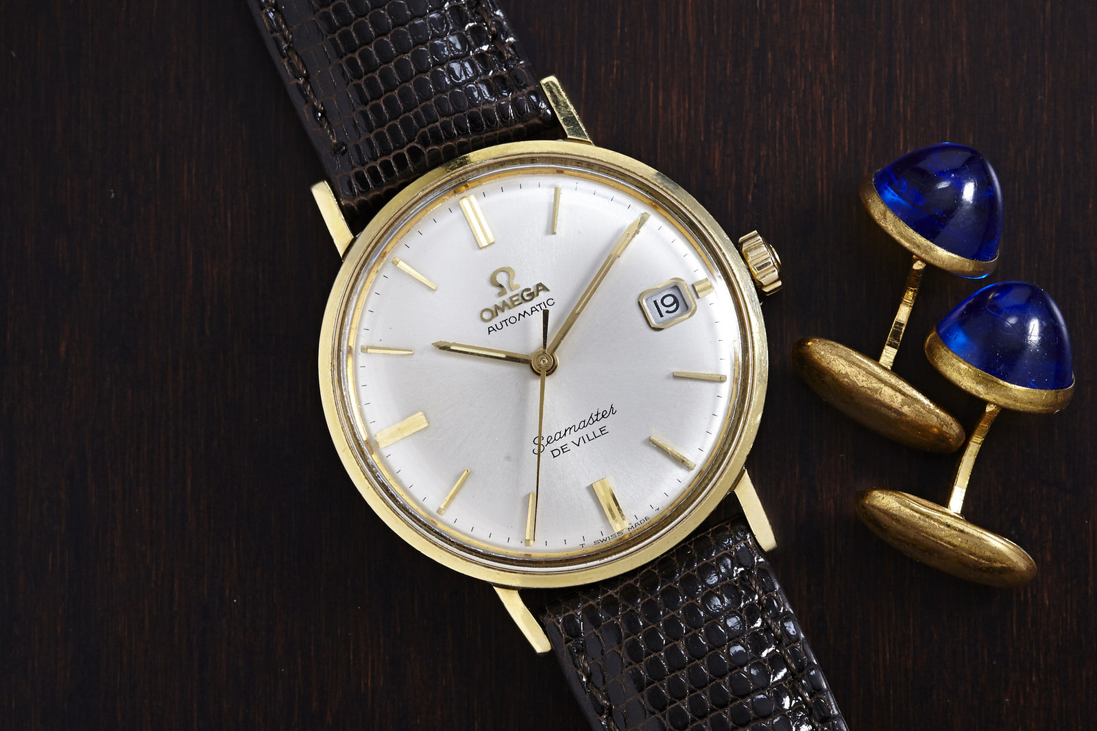 An Omega Seamaster De Ville Automatic wristwatch by Omega, featuring a gold case and brown leather strap, is shown on a dark wooden surface beside gold cufflinks with blue tops.