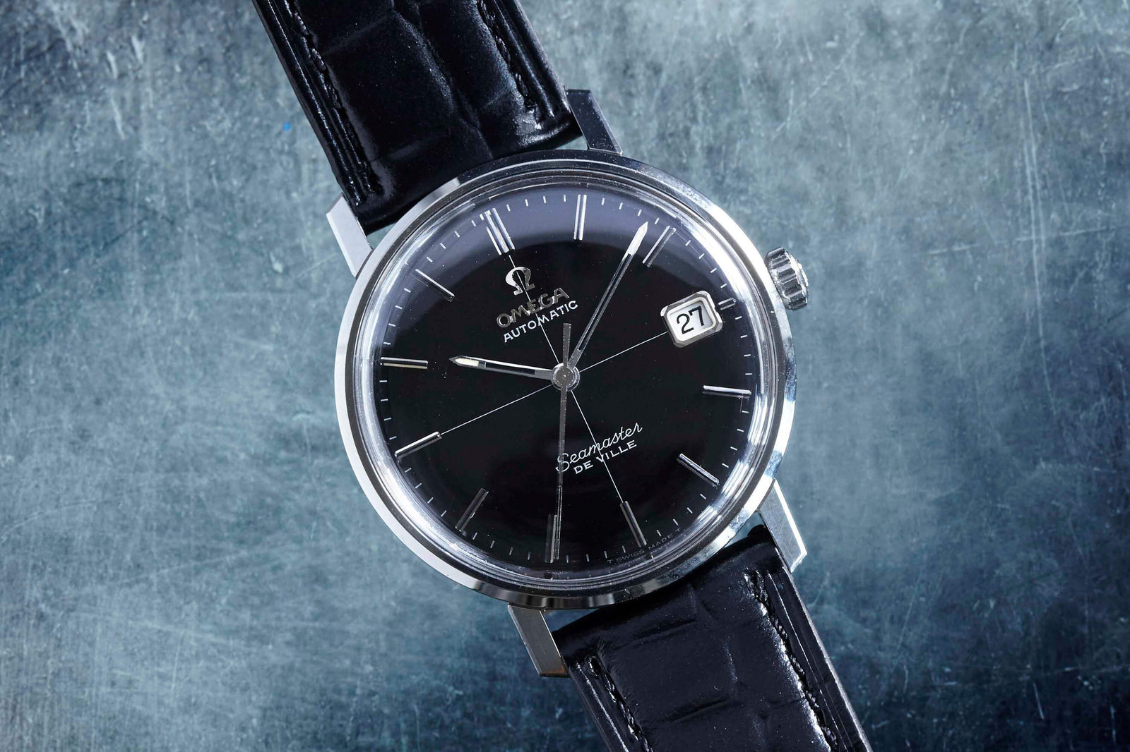 A close-up of the Omega Seamaster De Ville by Omega, a vintage black automatic wristwatch with leather strap, silver bezel, and date set to 27th, displayed on textured grey—an icon of classic men's style.