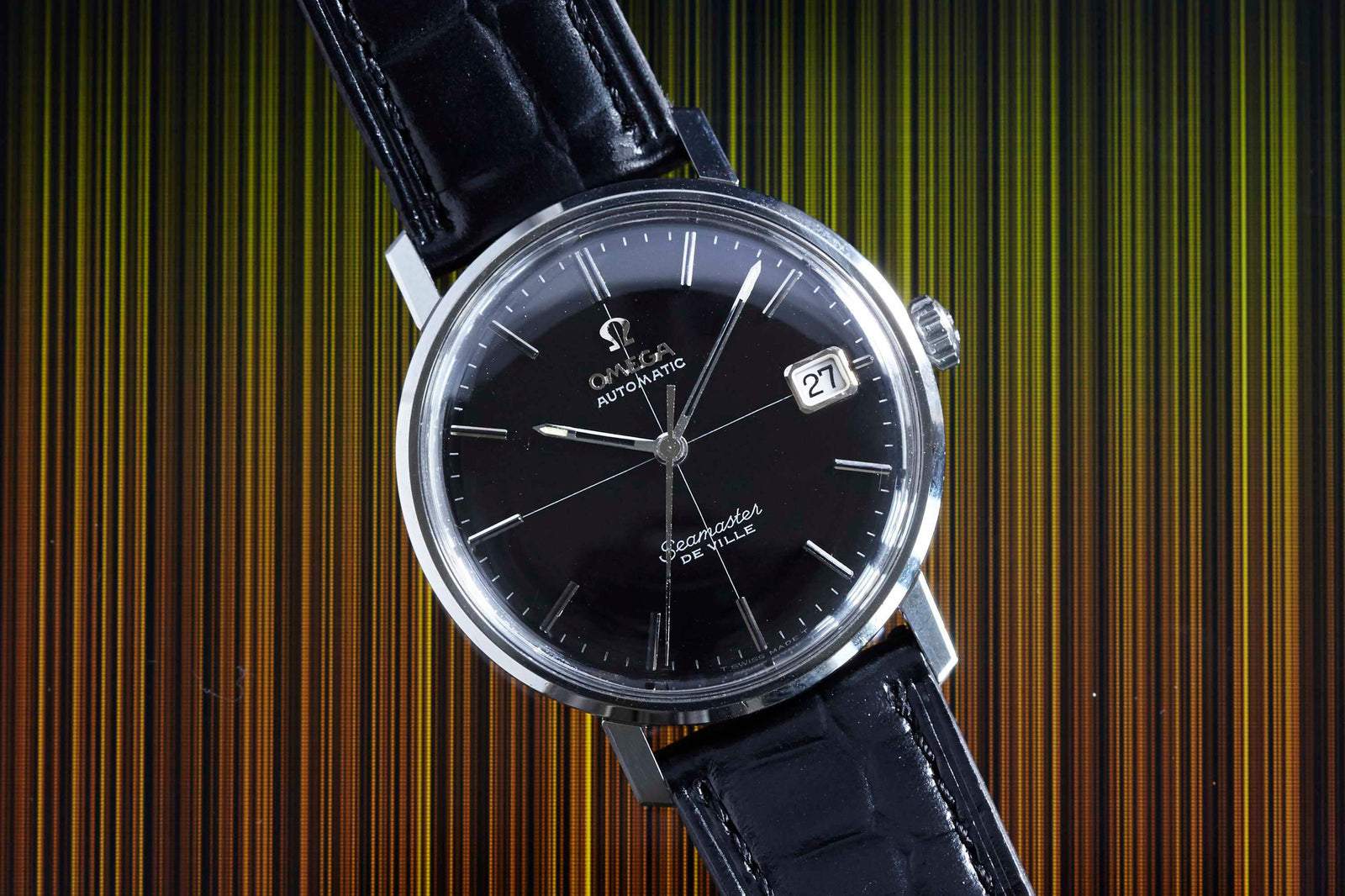 An Omega Seamaster De Ville automatic wristwatch with a black dial, silver markers, and a black leather strap is displayed against a striped background. The date window shows 27—a timeless example of classic men's style by Omega.