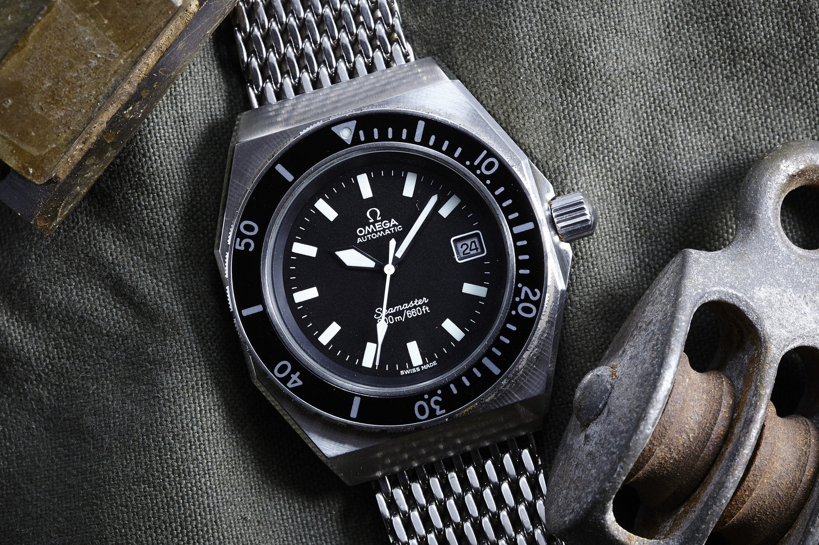 A close-up of the Omega Seamaster SHOM - Gear Patrol Exclusive, a vintage dive watch with a black dial and stainless steel mesh strap, rests on green fabric beside a metallic climbing pulley.
