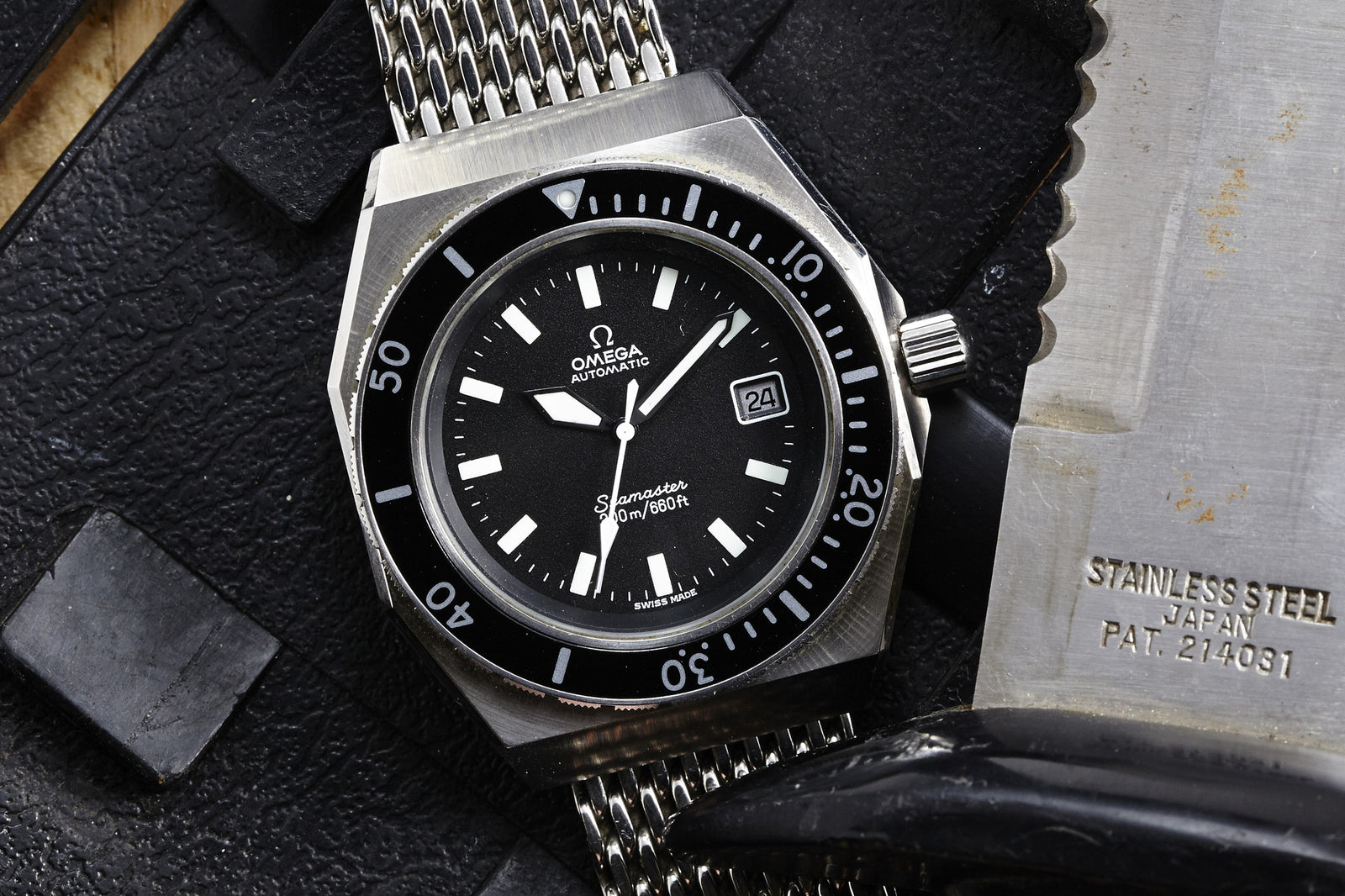 A close-up of the Omega Seamaster SHOM - Gear Patrol Exclusive by Omega, featuring a black dial and stainless steel mesh bracelet, displayed beside a stainless steel knife on a textured black surface.