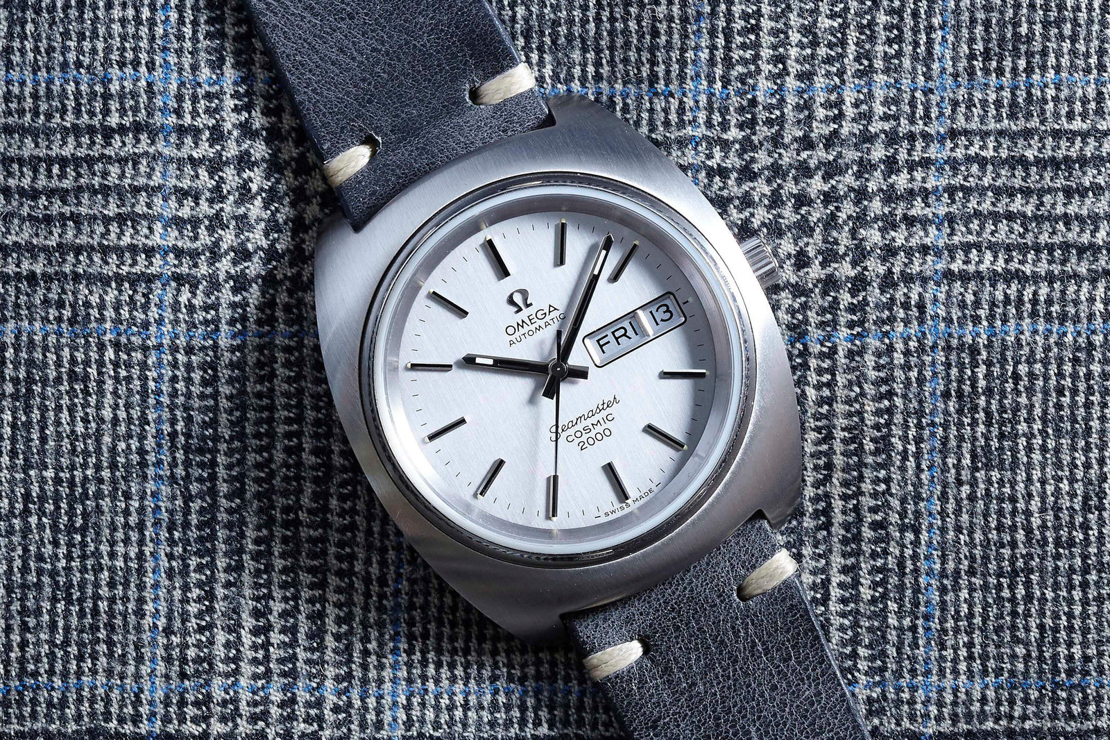An Omega Seamaster Cosmic by Omega with a white dial, black leather strap, and day-date display is displayed on a gray plaid fabric featuring blue and white lines.