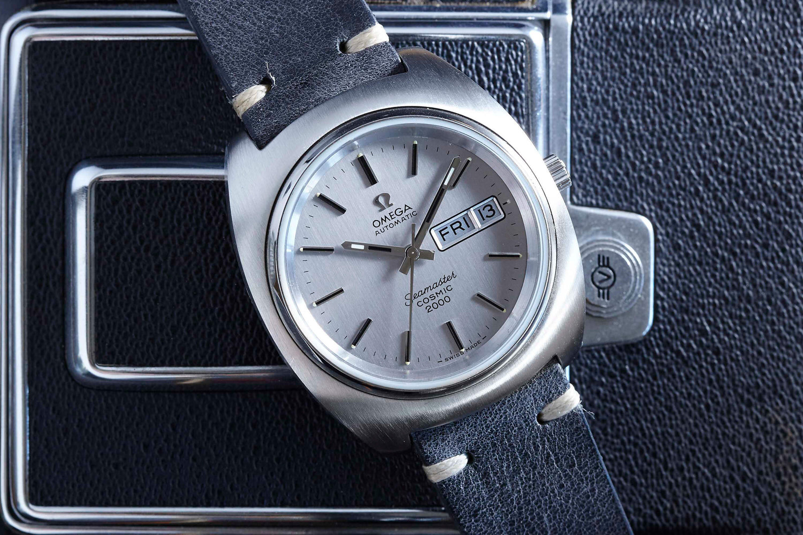 The Omega Seamaster Cosmic by Omega features a black leather strap and day-date display, shown here on Friday the 13th atop a textured black surface with metallic details—a must-have vintage piece for collectors.