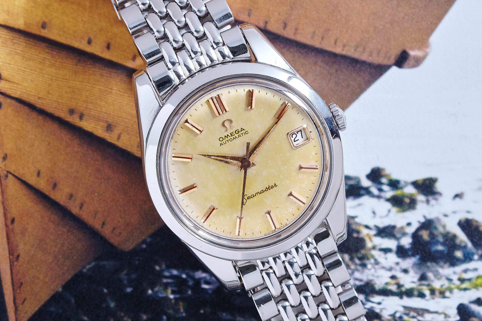 A close-up of the Omega Seamaster Automatic by Omega, featuring a steel case, cream dial, and date display at 3 o’clock, shown resting on a surface with wooden objects and pebbles in the background.