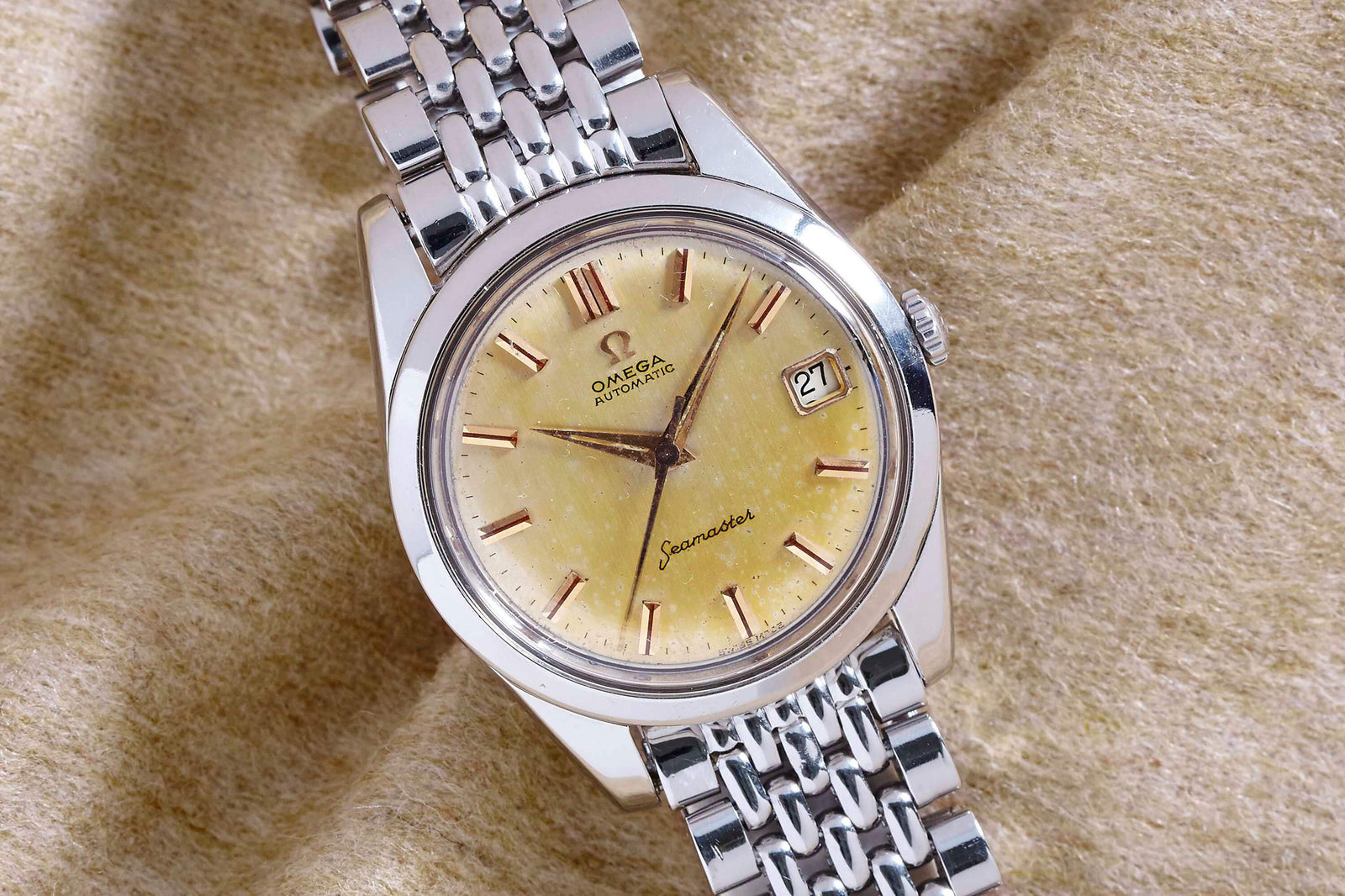 An Omega Seamaster Automatic wristwatch by Omega, featuring a yellow textured dial, gold hour markers, date at 3 o’clock, and a silver bracelet, rests on beige fabric—a collectible vintage automatic steel timepiece.
