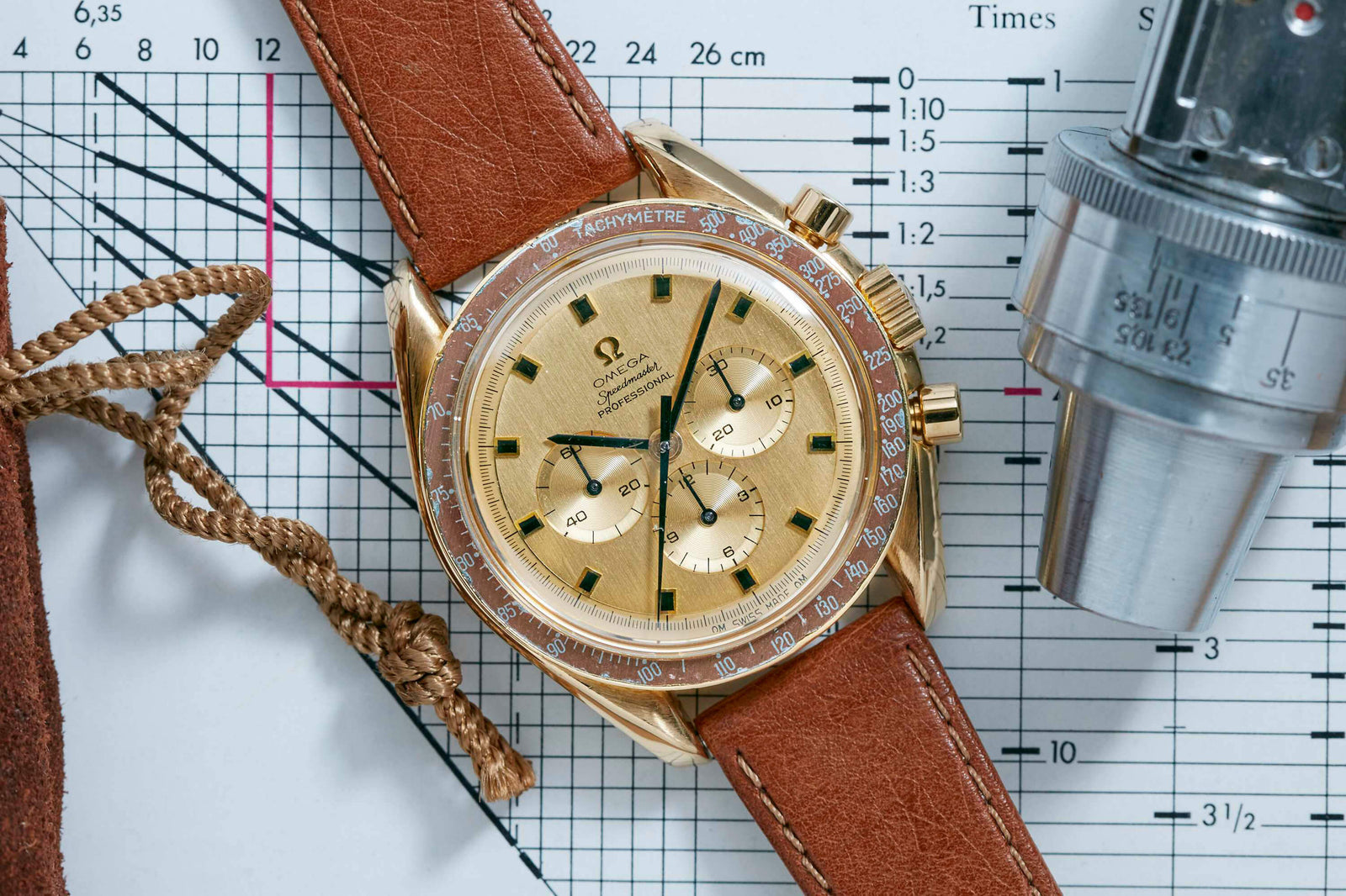 An Omega Speedmaster Professional 18k Gold with a brown leather strap sits on technical graph paper beside a metal tool and coiled brown cord. This iconic Omega watch features three subdials and a tachymeter bezel.