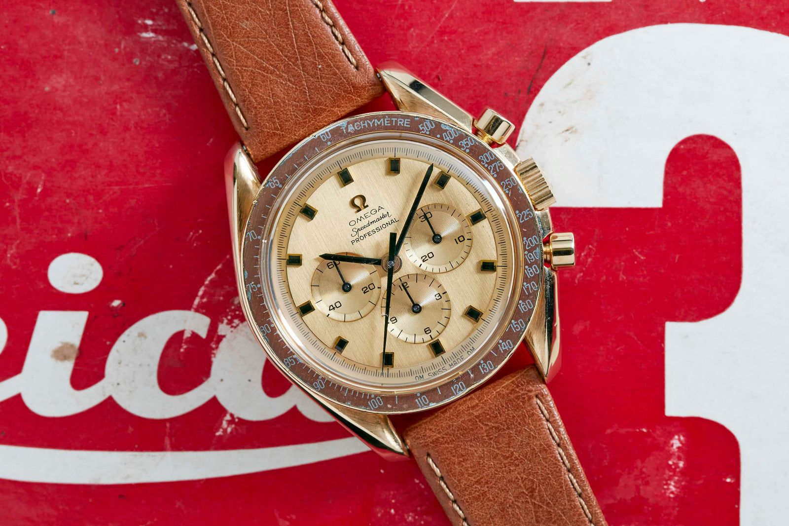 An Omega Speedmaster Professional 18k Gold chronograph wristwatch with a brown leather strap is set on a red and white background, featuring three sub-dials and a tachymeter bezel. This iconic Omega watch is linked to NASA space missions.