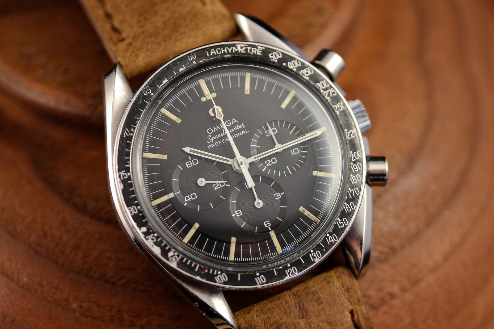 A close-up of the Omega Speedmaster - 1968 Transitional Dial wristwatch by Omega, featuring a black dial with three subdials, a tachymeter bezel, and brown leather strap, elegantly displayed on a wooden surface.