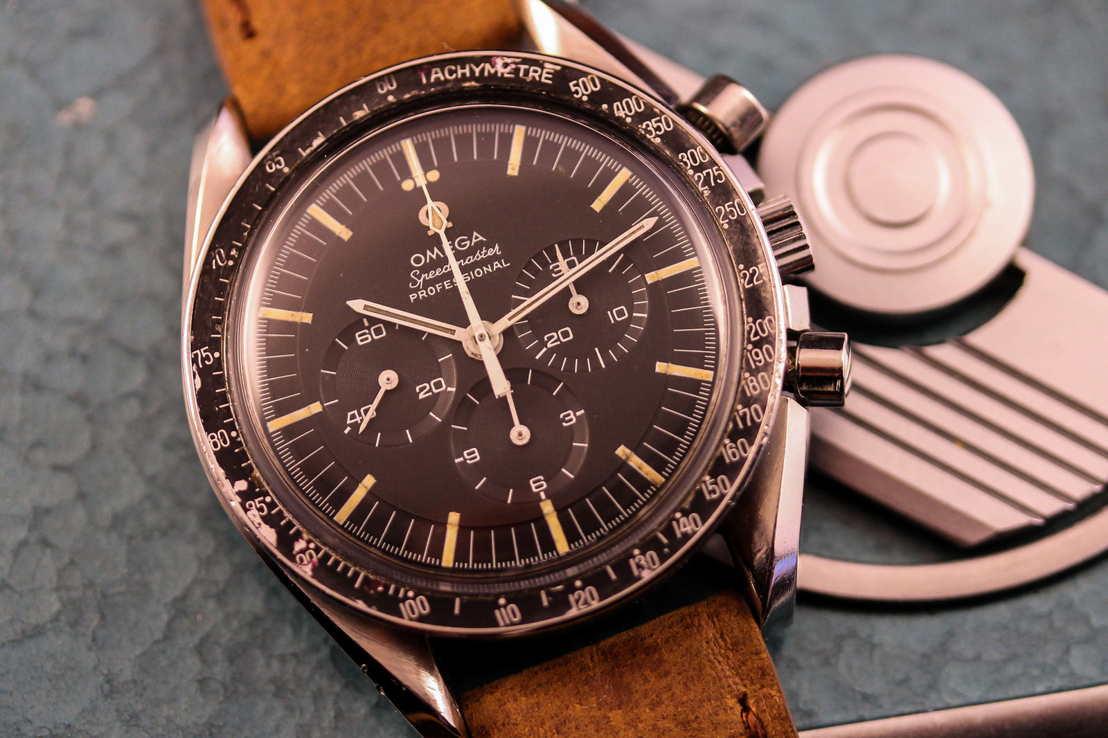 A close-up showcases the Omega Speedmaster - 1968 Transitional Dial by Omega, featuring a black dial with chronograph subdials and a brown leather strap, set on a textured blue surface beside a metallic object.