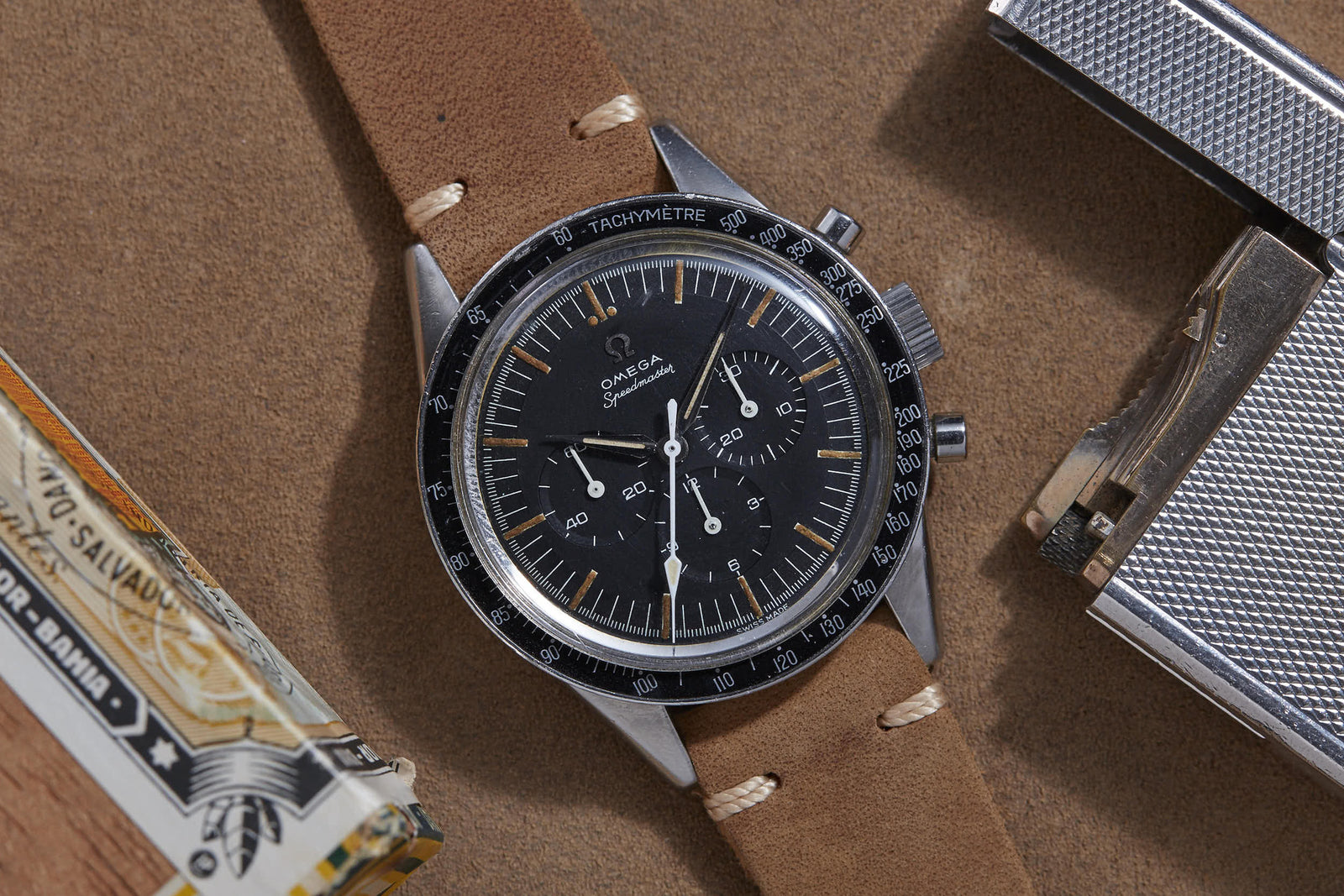 An Omega Speedmaster by Omega with a brown leather strap, black dial, and tachymeter bezel rests atop a brown surface beside a metallic lighter and cigar box—evoking the style of iconic Speedmaster auctions.