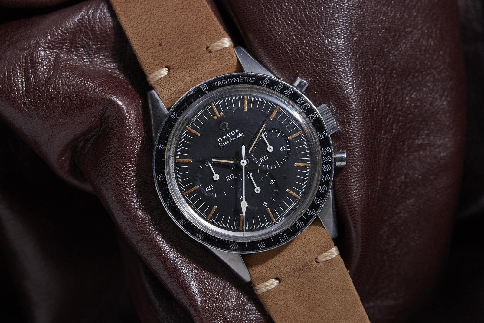 An Omega Speedmaster wristwatch by Omega, featuring a black dial, chronograph subdials, and a brown leather strap, rests elegantly on dark brown leather fabric.