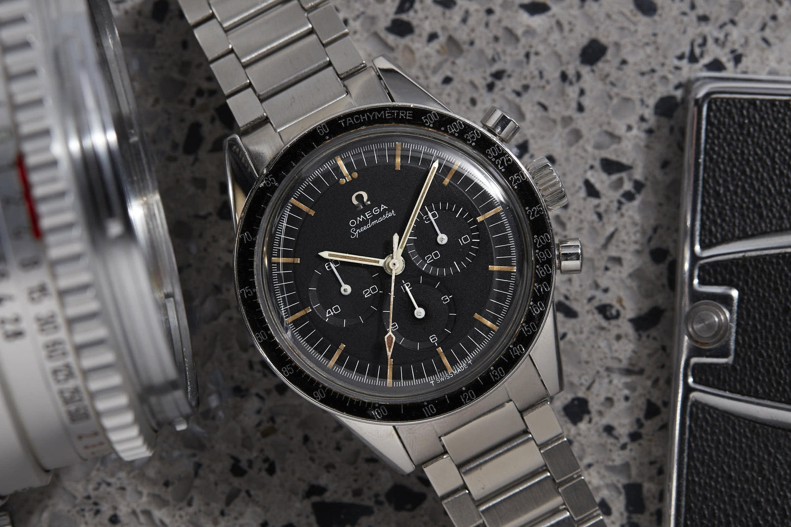 A close-up of the Omega Speedmaster "Ed White" by Omega, featuring a stainless steel bracelet and linked to the Gemini IV mission, rests on a speckled surface beside a camera lens and a black object.