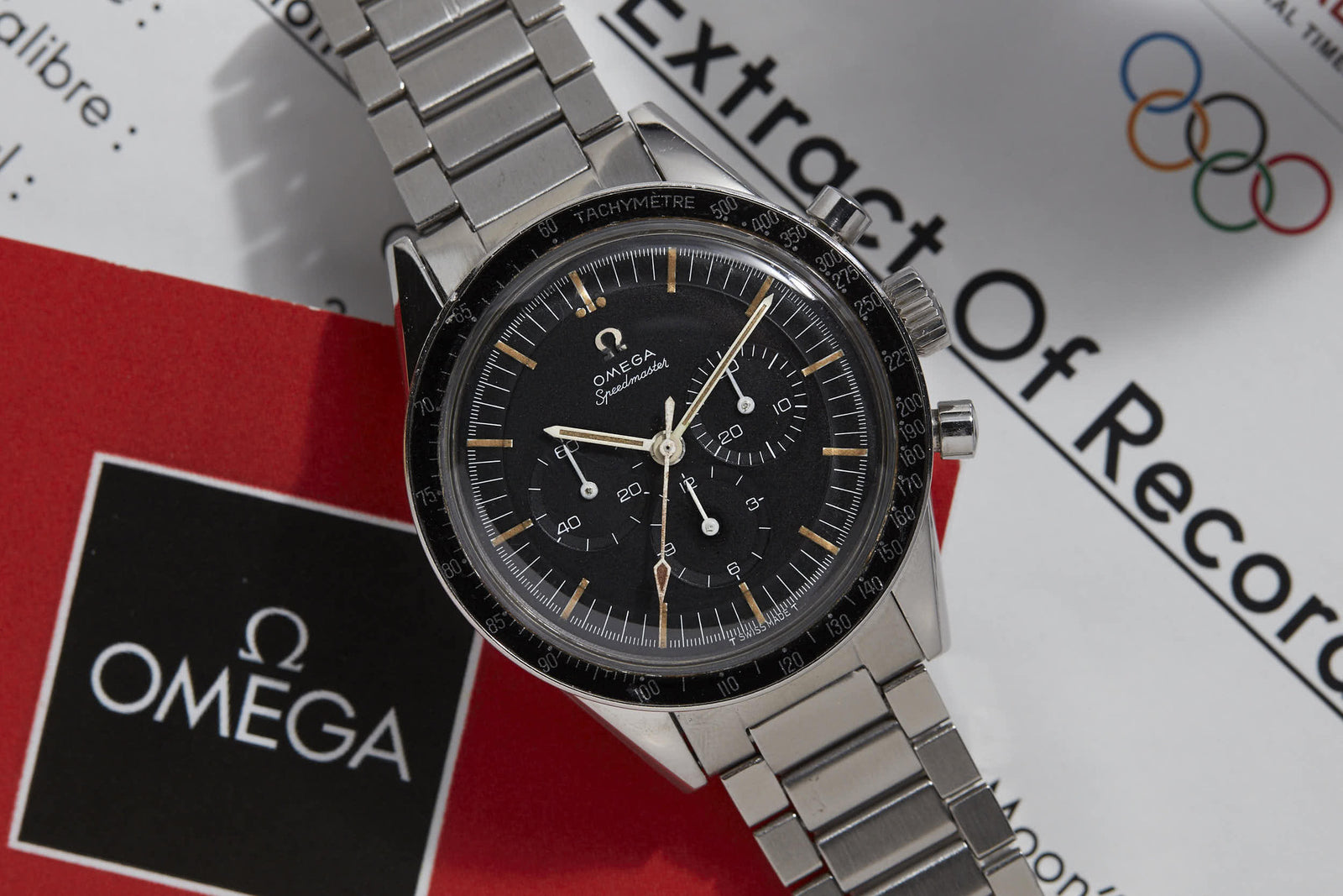 A close-up of the Omega Speedmaster "Ed White" by Omega with a stainless steel band, resting on an Omega booklet and an Extract Of Records document featuring Olympic rings—highlighting its NASA Gemini IV mission heritage.