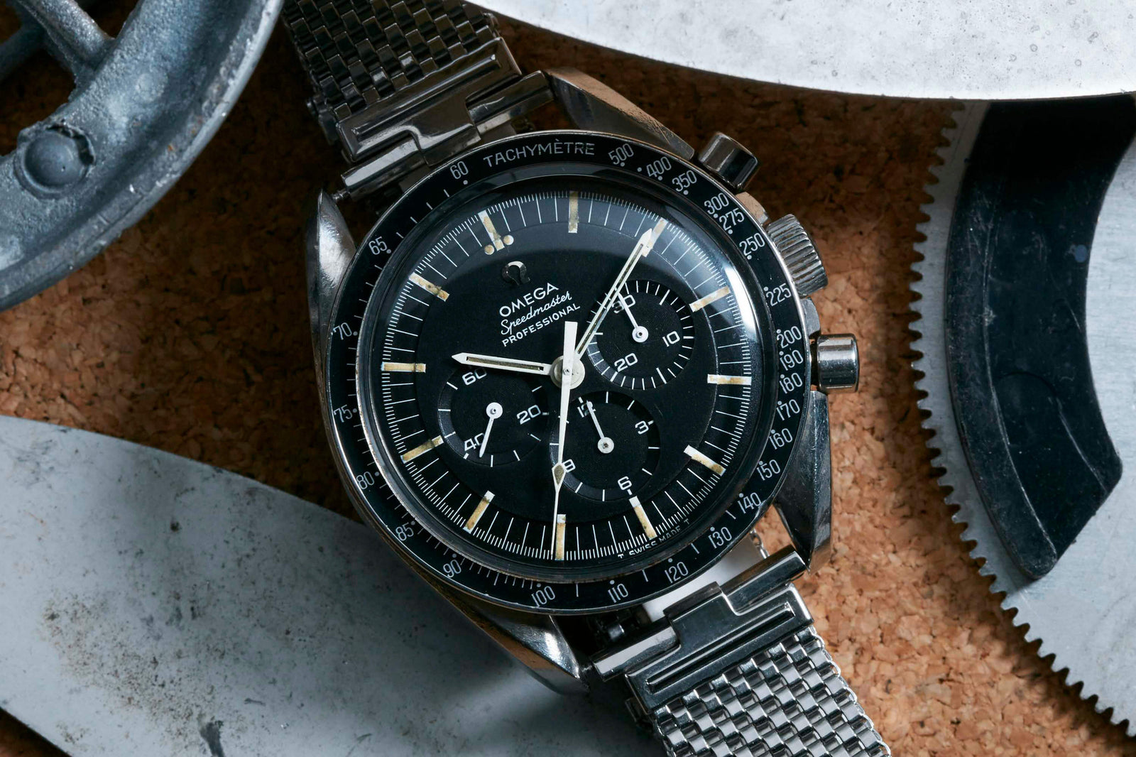 A close-up of the Omega Speedmaster Professional - 1967 by Omega, featuring a black dial, tachymeter bezel, and metal mesh strap on cork among gears—a legendary watch worn during Apollo missions and the historic moon landing.