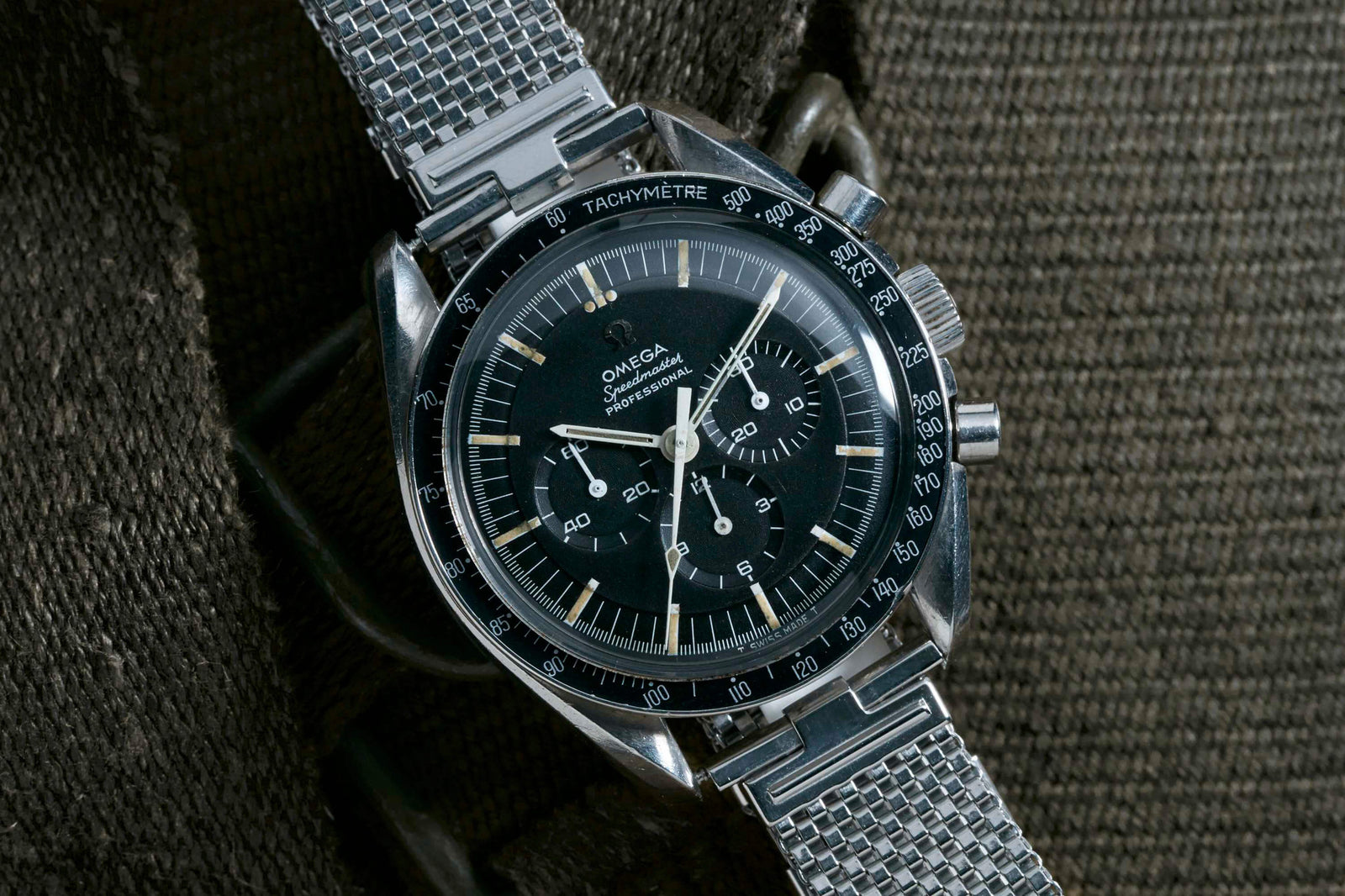 A close-up of the Omega Speedmaster Professional - 1967 by Omega, featuring a black dial and silver mesh strap, rests on textured brown fabric. This iconic model, worn during Apollo missions, shows the time and multiple subdials.