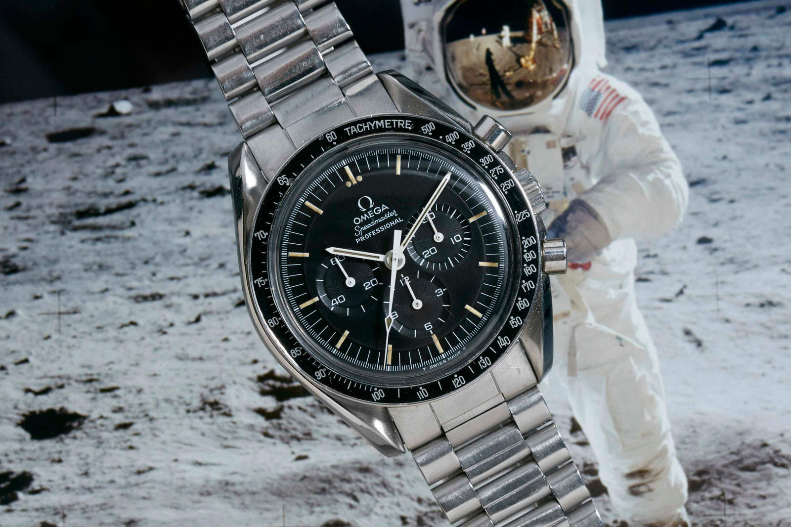 A close-up of the Omega Speedmaster Professional - 1969 by Omega highlights its stainless steel case and black dial, with a blurred background showing an astronaut on the moon’s surface in a spacesuit.