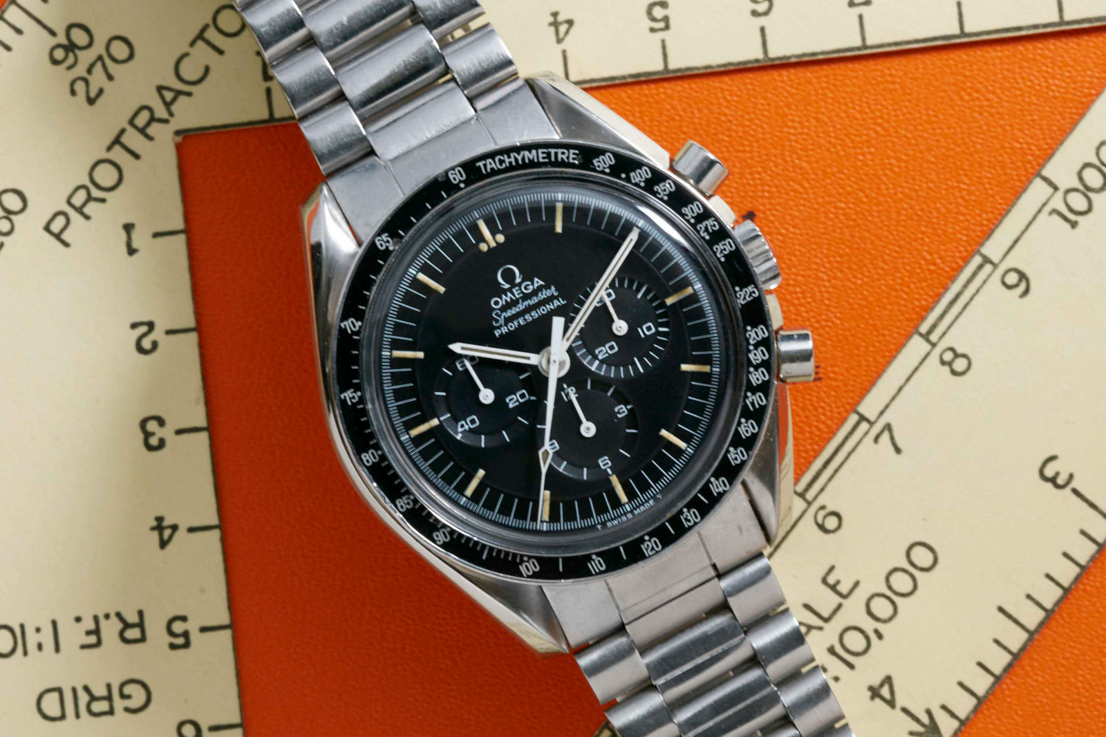 An Omega Speedmaster Professional - 1969 by Omega, with a black dial and tachymeter bezel, is displayed on orange and cream geometric papers marked with measurement lines and protractor scales.