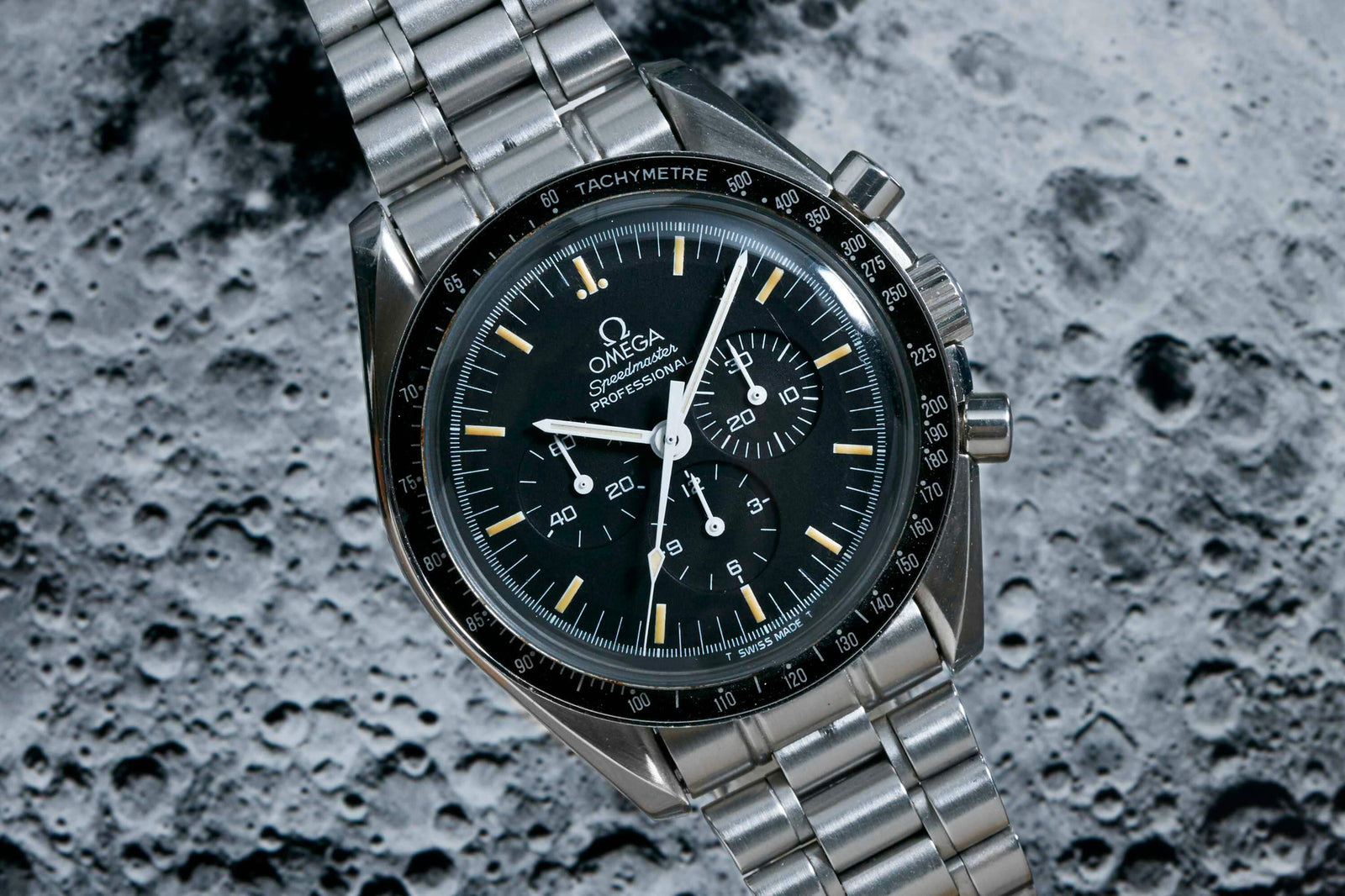 The Omega Speedmaster Professional - 1993 by Omega features a stainless steel case, black dial, chronograph functions, and matching metal bracelet, showcased against a textured surface reminiscent of the moon.
