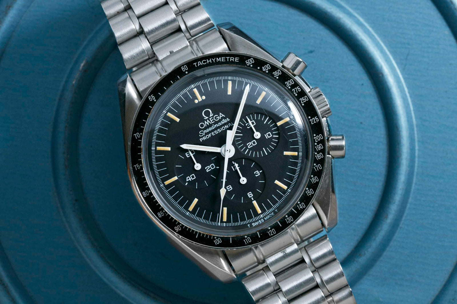 The Omega Speedmaster Professional - 1993 by Omega, with a black dial, three subdials, and stainless steel bracelet, channels Apollo-era style as it rests on a blue textured surface.