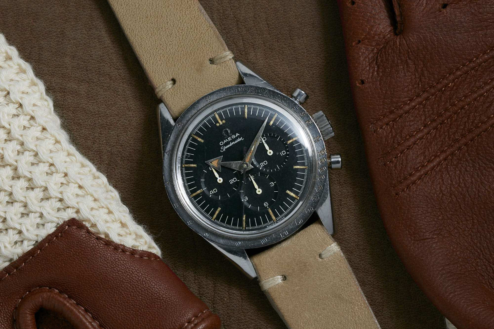 An Omega Speedmaster racing chronograph by Omega with a beige leather strap rests on a brown surface, alongside brown leather gloves and cream knit fabric.