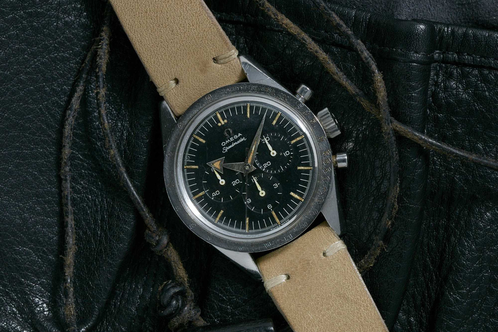 An Omega Speedmaster by Omega with a black dial, beige indices, and a tan leather strap rests elegantly on a black leather surface.
