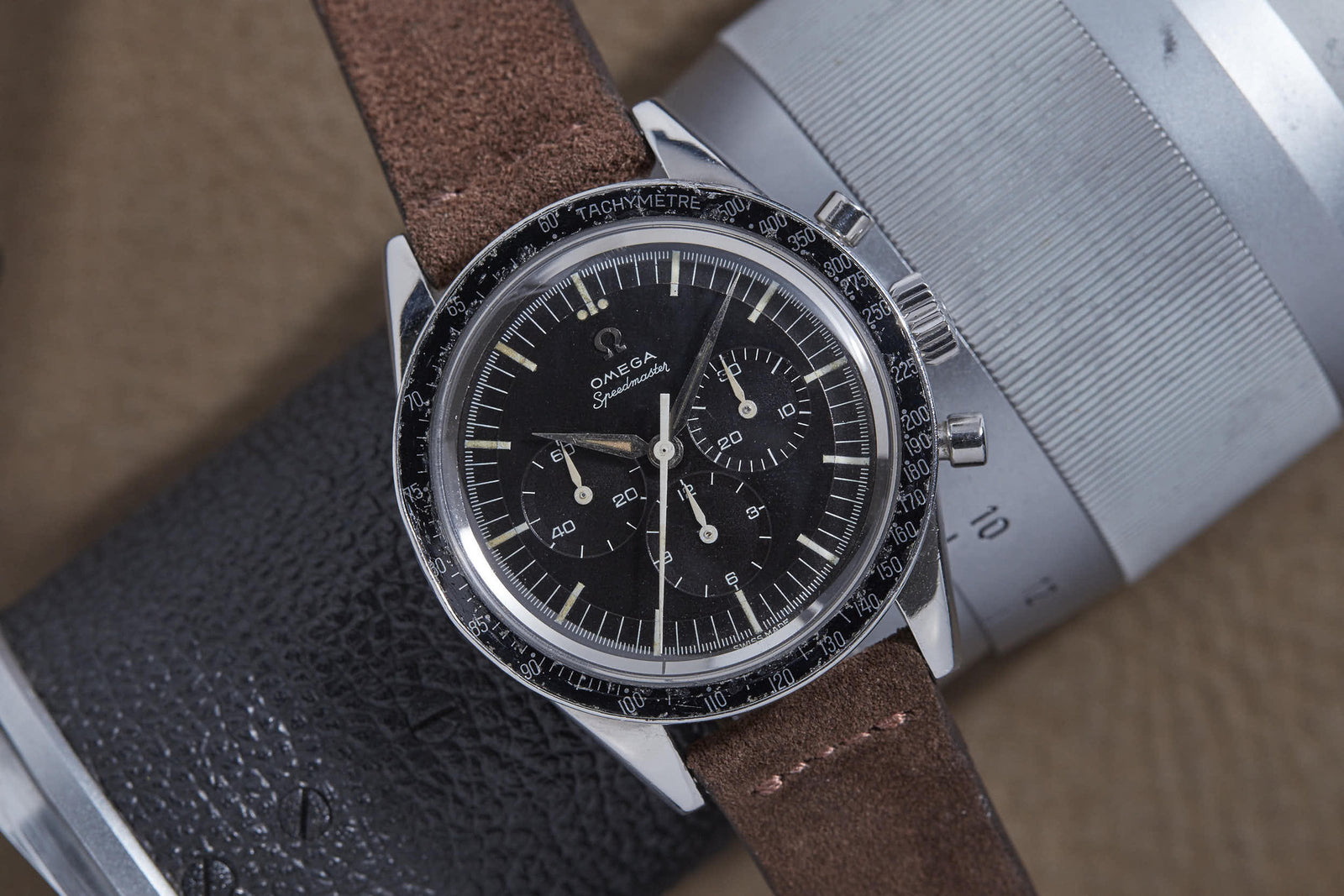 An Omega Speedmaster Ref. 2998-3 wristwatch by Omega, known for its Moon landing heritage, features a black dial and brown leather strap, resting on a textured black and silver cylindrical display.