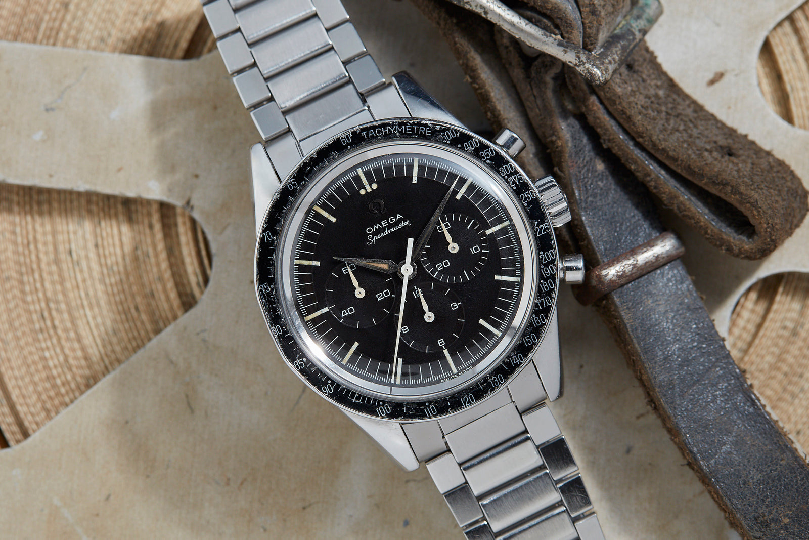 An Omega Speedmaster Ref. 2998-3 stainless steel watch with black dial and tachymeter bezel sits on a metal bracelet next to a worn brown leather strap on a light textured surface.