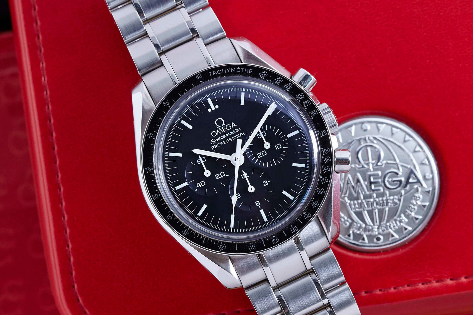 An Omega Speedmaster stainless steel chronograph wristwatch with a black dial is shown on a red Omega case beside a silver Omega medallion—reflecting the Apollo-era legacy and modern design of this iconic Omega timepiece.