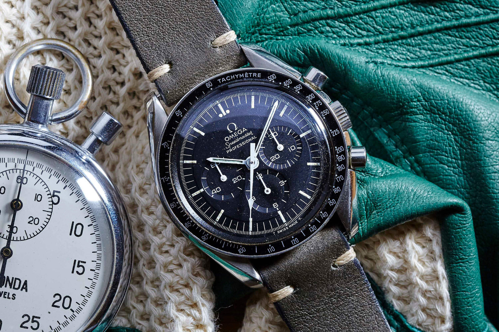 An Omega Speedmaster Professional - 1969 chronograph with a black dial and tachymeter bezel rests on a green leather glove, beside a silver stopwatch and cream knit fabric.