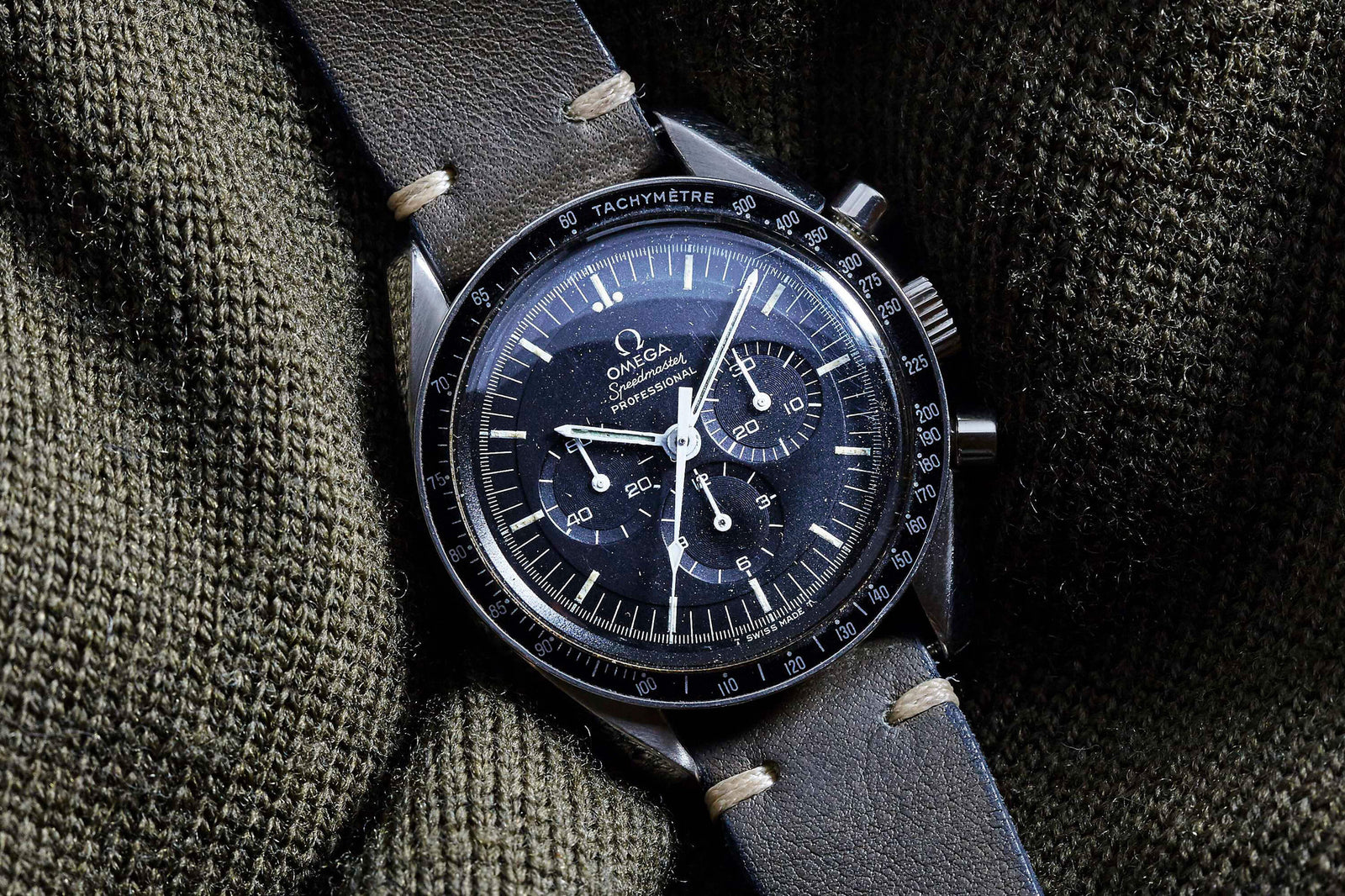 A close-up of the Omega Speedmaster Professional - 1969 by Omega, featuring a black dial, tachymeter bezel, and vintage chronograph design, displayed on dark green fabric with a black leather strap and beige stitching.