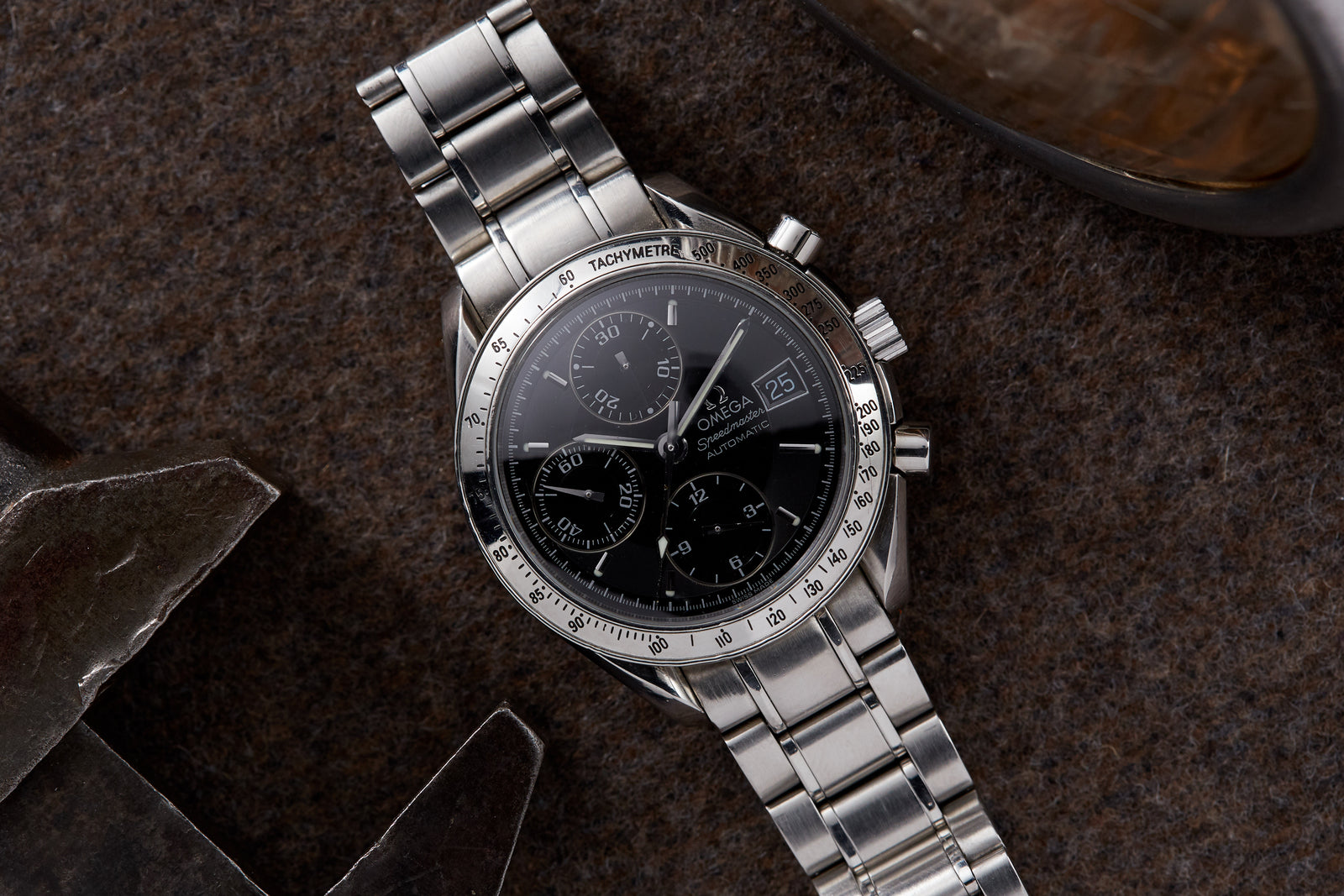An Omega Speedmaster Reduced Automatic Date wristwatch with a black dial and tachymeter bezel rests on a brown textured surface near a metal object.