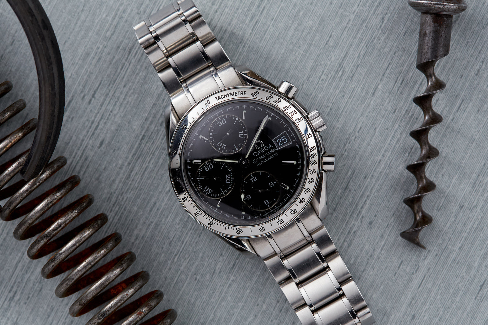 An Omega Speedmaster Reduced Automatic Date watch with a black dial and chronograph sits on a grey surface, surrounded by metal objects—a modern stainless steel timepiece from Omega.