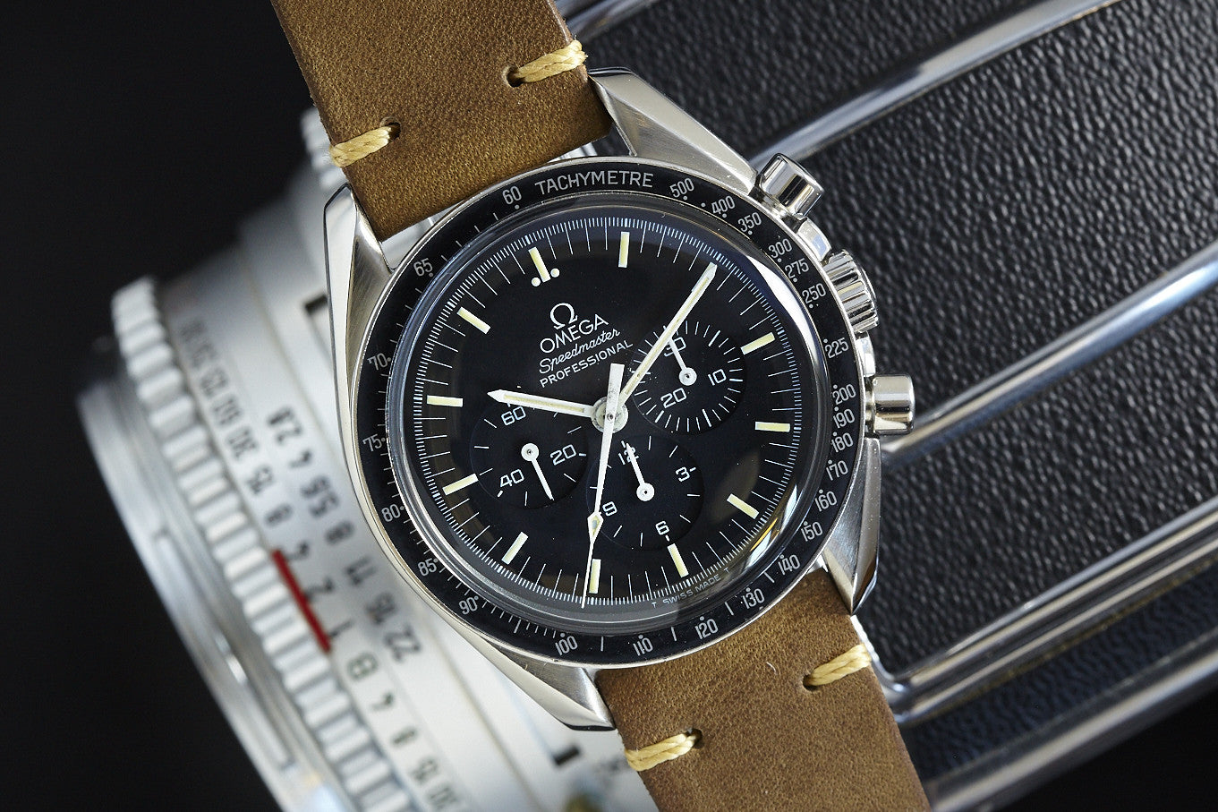 An Omega Speedmaster by Omega, featuring a black dial and tan leather strap, is placed on a silver camera lens marked with numbers.