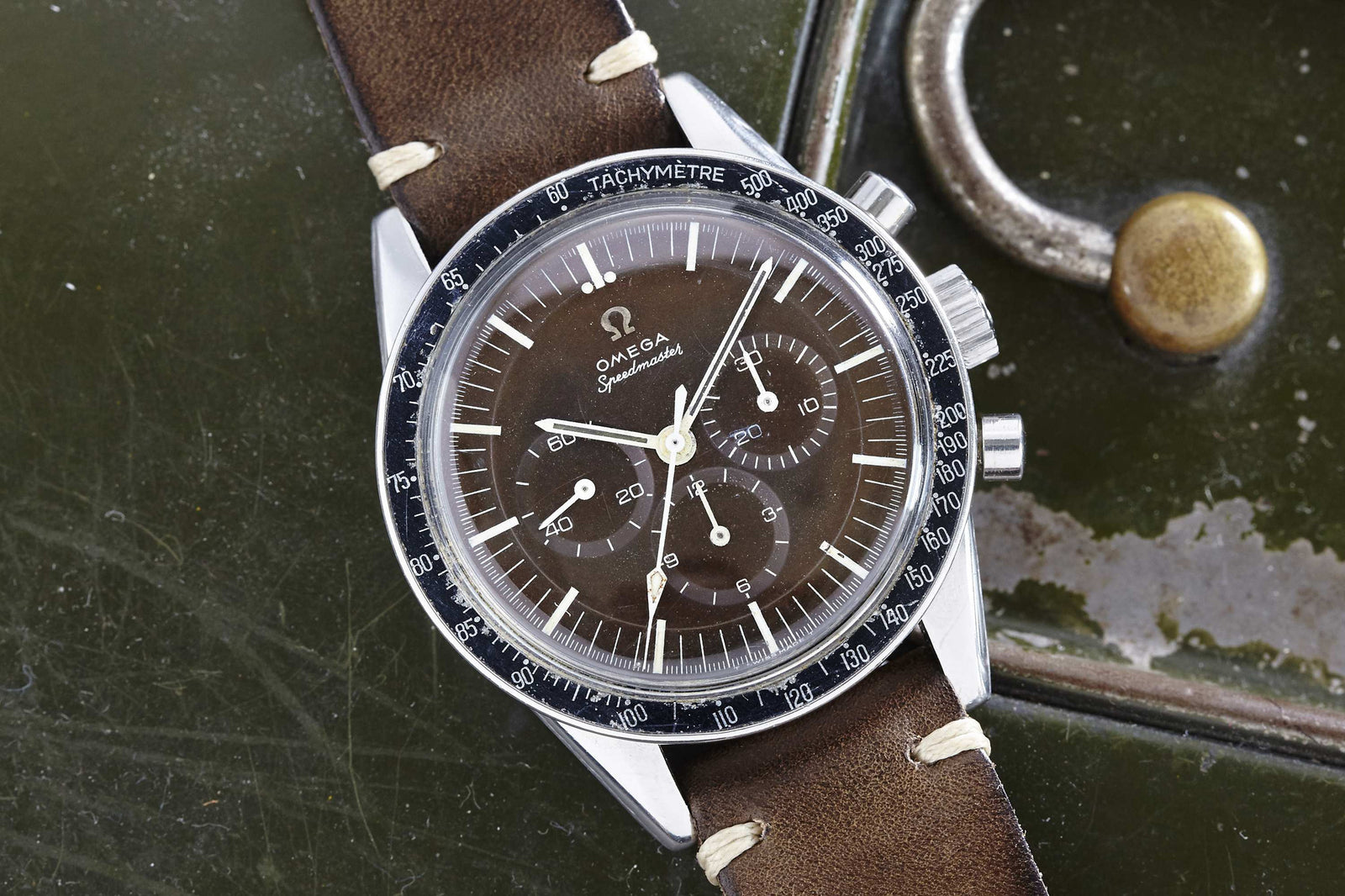 A close-up of an Omega Speedmaster Ed White watch with a brown dial, white markers, silver case, and brown leather strap, placed on a green metallic surface with a brass fitting.