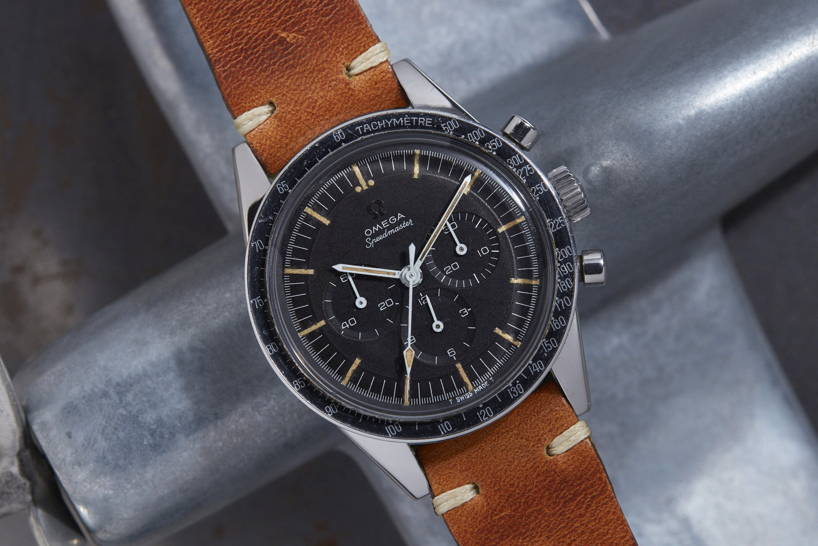 The Omega Speedmaster "Ed White" by Omega features a black dial with three subdials and a brown leather strap, showcased on a metallic surface—a historic watch celebrated for its connection to NASA missions.