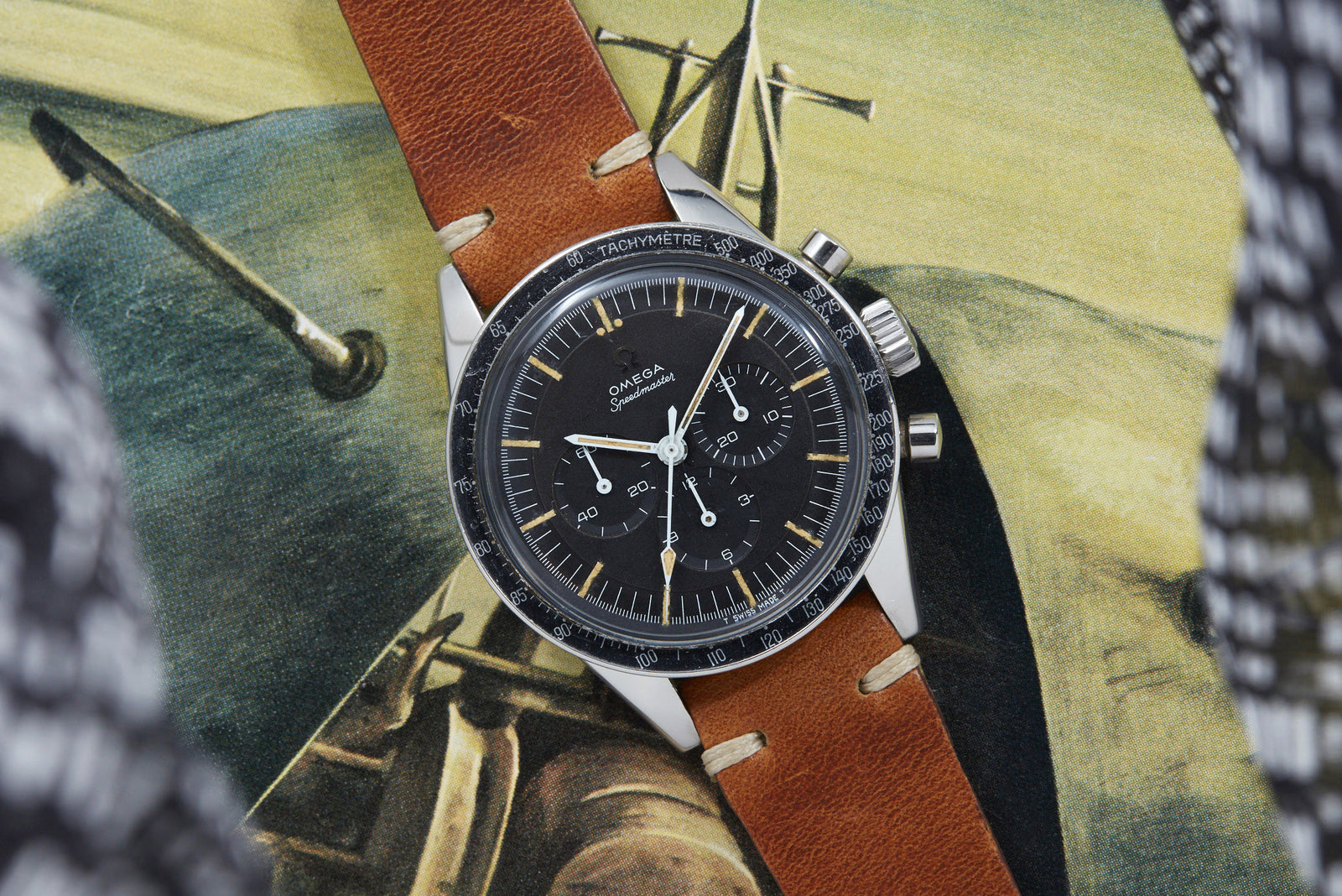 An Omega Speedmaster "Ed White" wristwatch by Omega with a brown leather strap rests on an aviation-themed painting featuring aircraft and a propeller, subtly referencing NASA history and partially framed by patterned fabric.