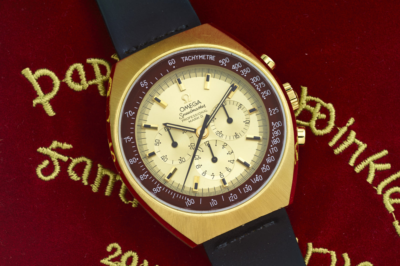 An Omega Speedmaster Mark II by Omega with a black strap is showcased on red velvet adorned with gold embroidered text in multiple scripts.