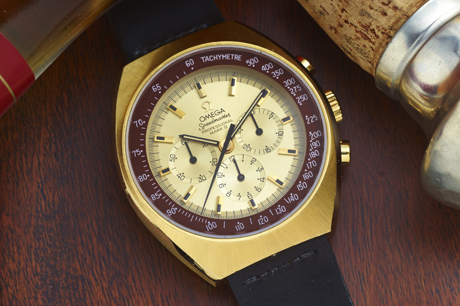 An Omega Speedmaster Mark II sports watch with a cream dial, black subdials, and a brown bezel featuring a tachymeter scale rests on a wooden surface beside metallic and cork objects.