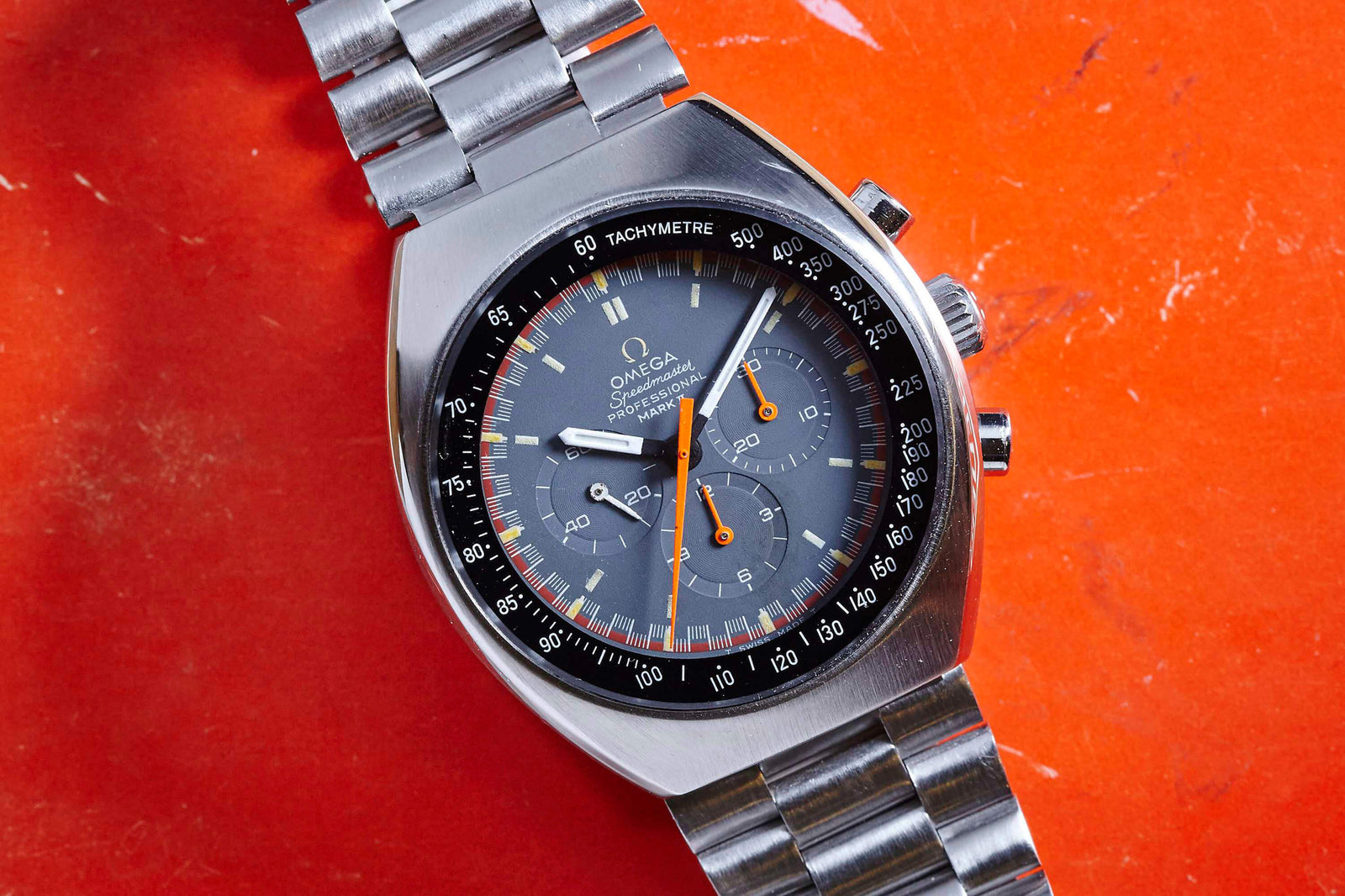 An Omega Speedmaster Mark II stainless steel wristwatch with a gray dial, orange accents, and tachymeter scale from Omega is displayed on a red textured surface.