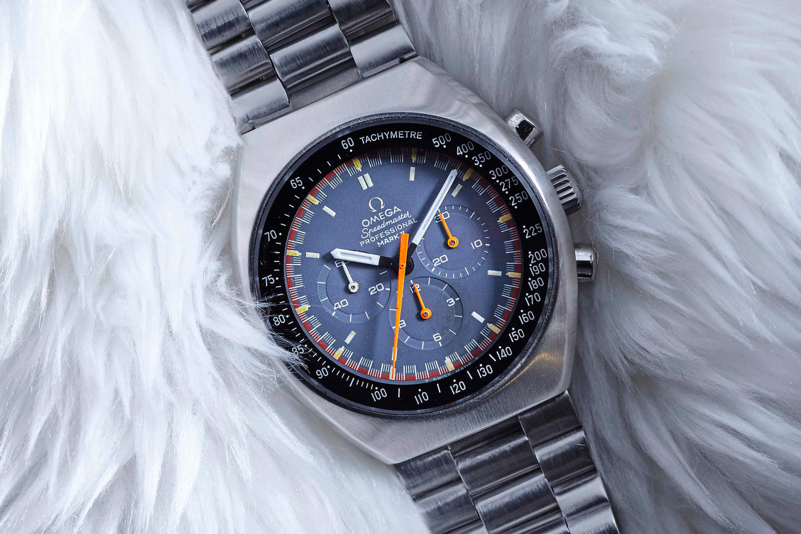 An Omega Speedmaster Mark II wristwatch with a blue dial, orange hands, and a tachymeter bezel by Omega rests on a soft, white surface.
