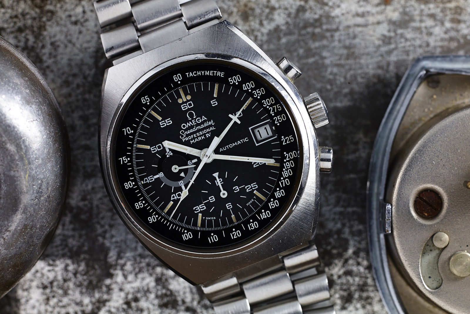 An Omega Speedmaster Mark IV Automatic stainless steel chronograph wristwatch with a black dial, three subdials, and date window is displayed on a textured metallic surface beside a partially visible metal object.