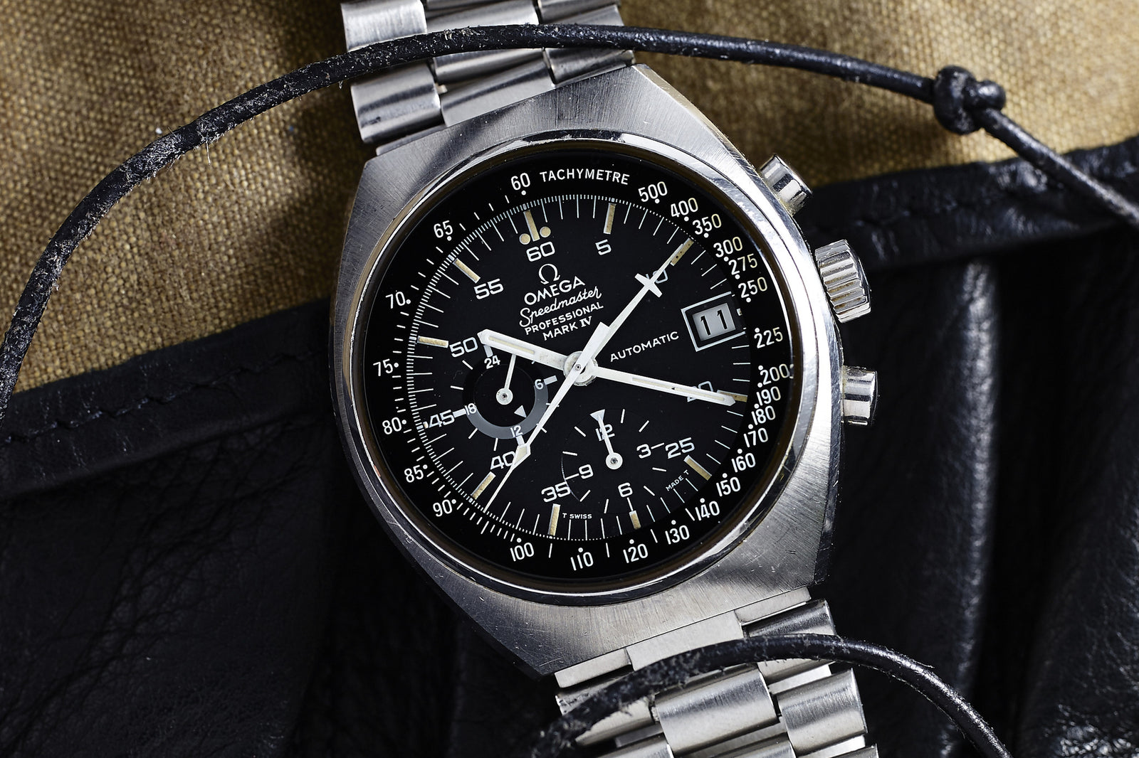 A close-up of the Omega Speedmaster Mark IV Automatic by Omega, featuring a stainless steel case, black dial, tachymeter bezel, and metal bracelet, resting on leather and fabric with a black cord draped over it.