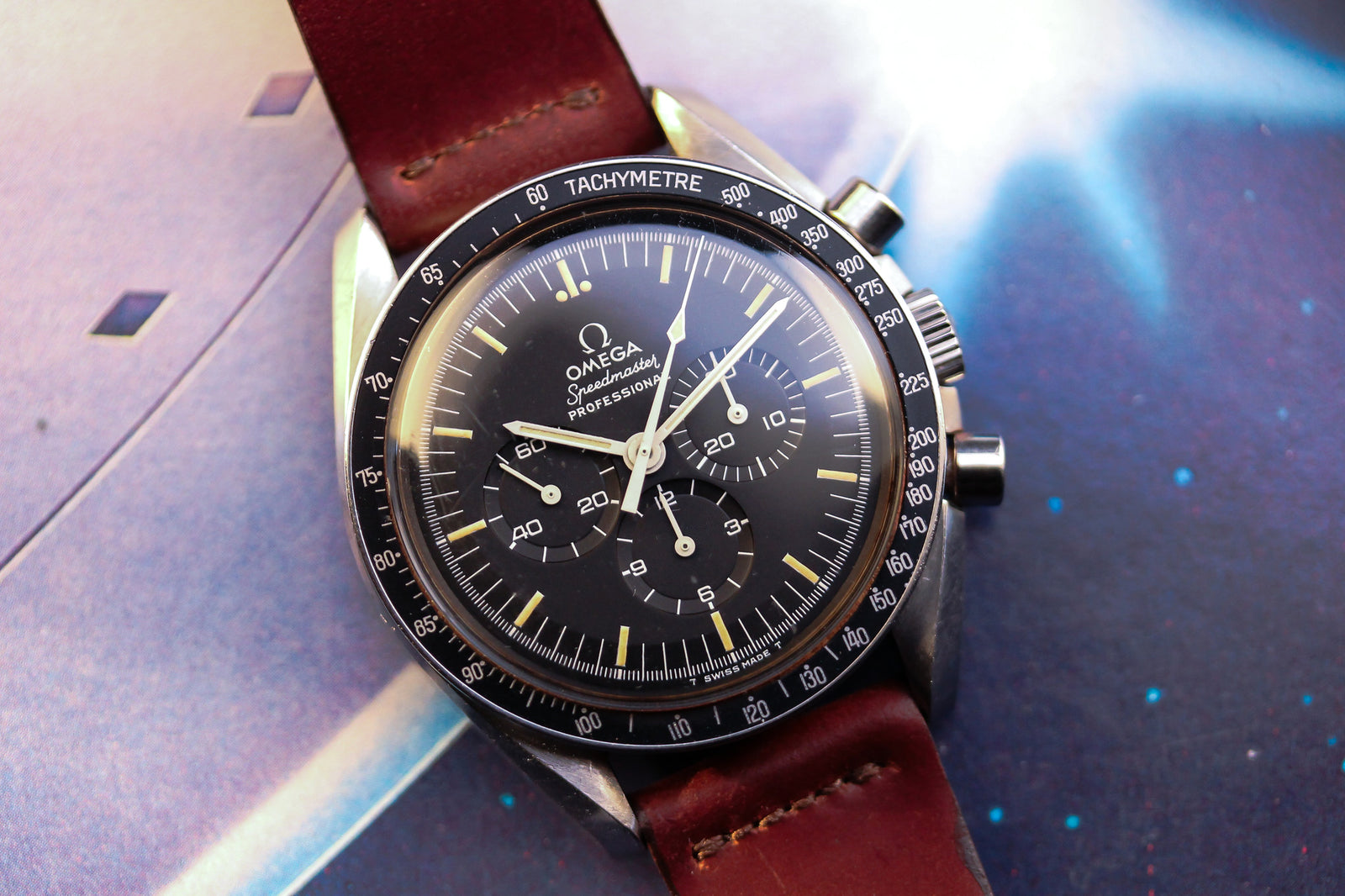 A close-up of the Omega Speedmaster Professional - 1974 by Omega, featuring a brown leather strap, black dial with three subdials and white markings, set against a blue background.