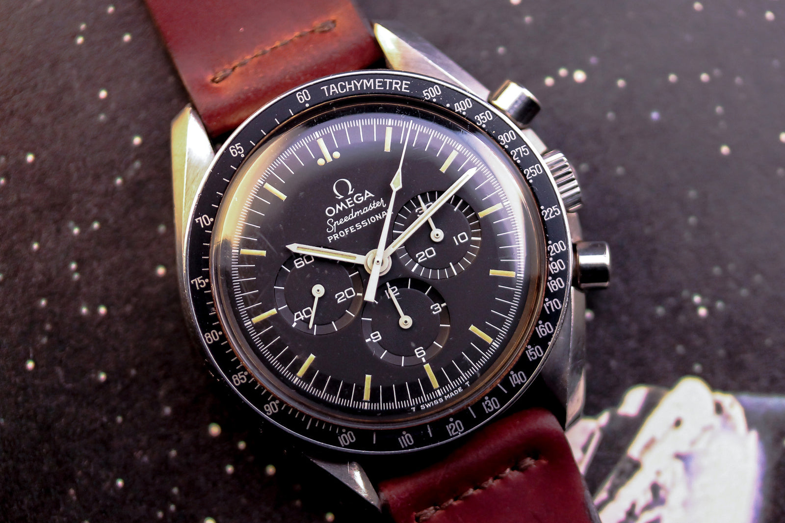 A close-up of the Omega Speedmaster Professional - 1974 by Omega, featuring a black dial with chronograph subdials, tachymeter bezel, and brown leather strap, set against a starry background.