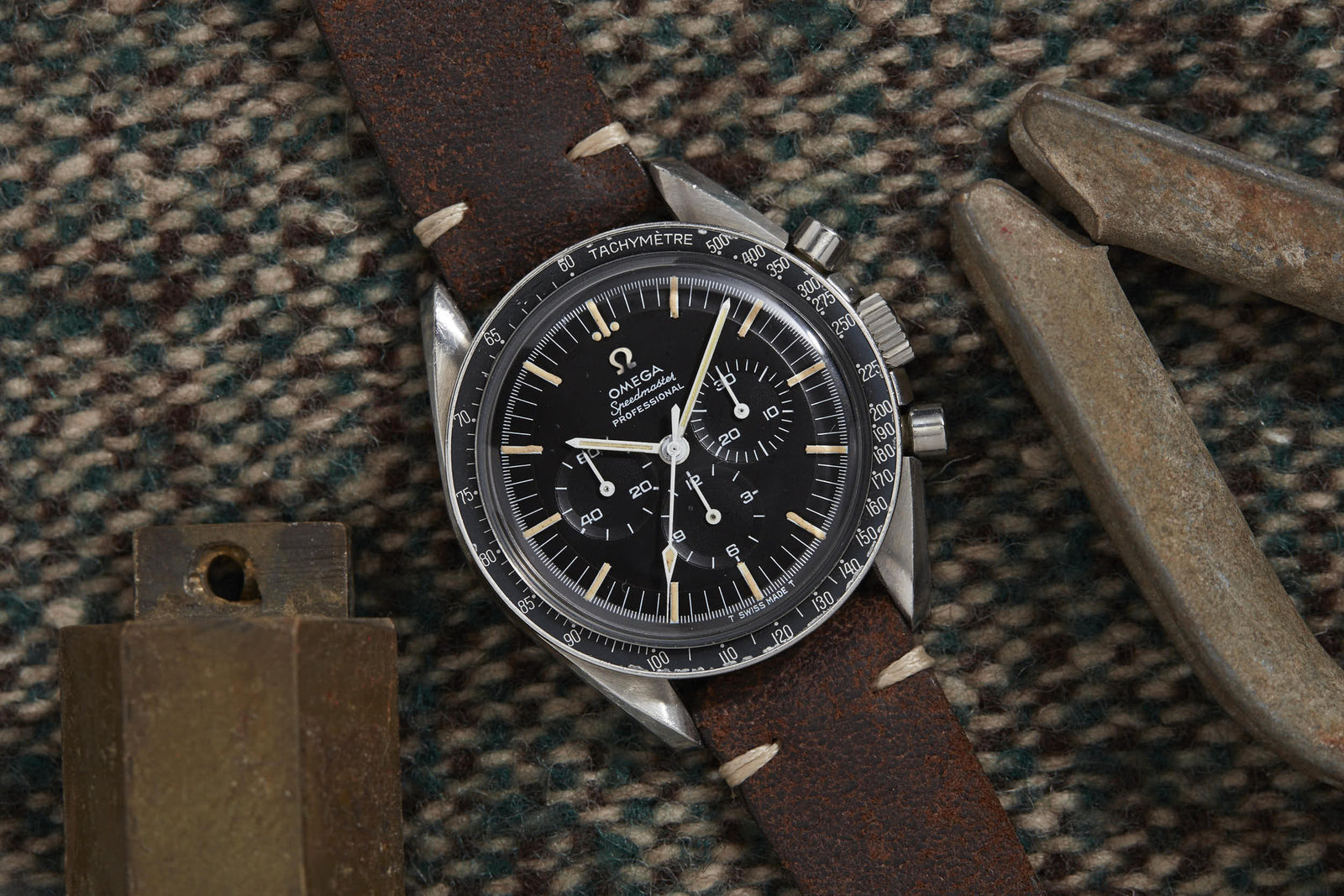 An Omega Speedmaster Professional by Omega with a dark face and brown leather strap rests on plaid wool fabric beside a brass object and a metal tool.