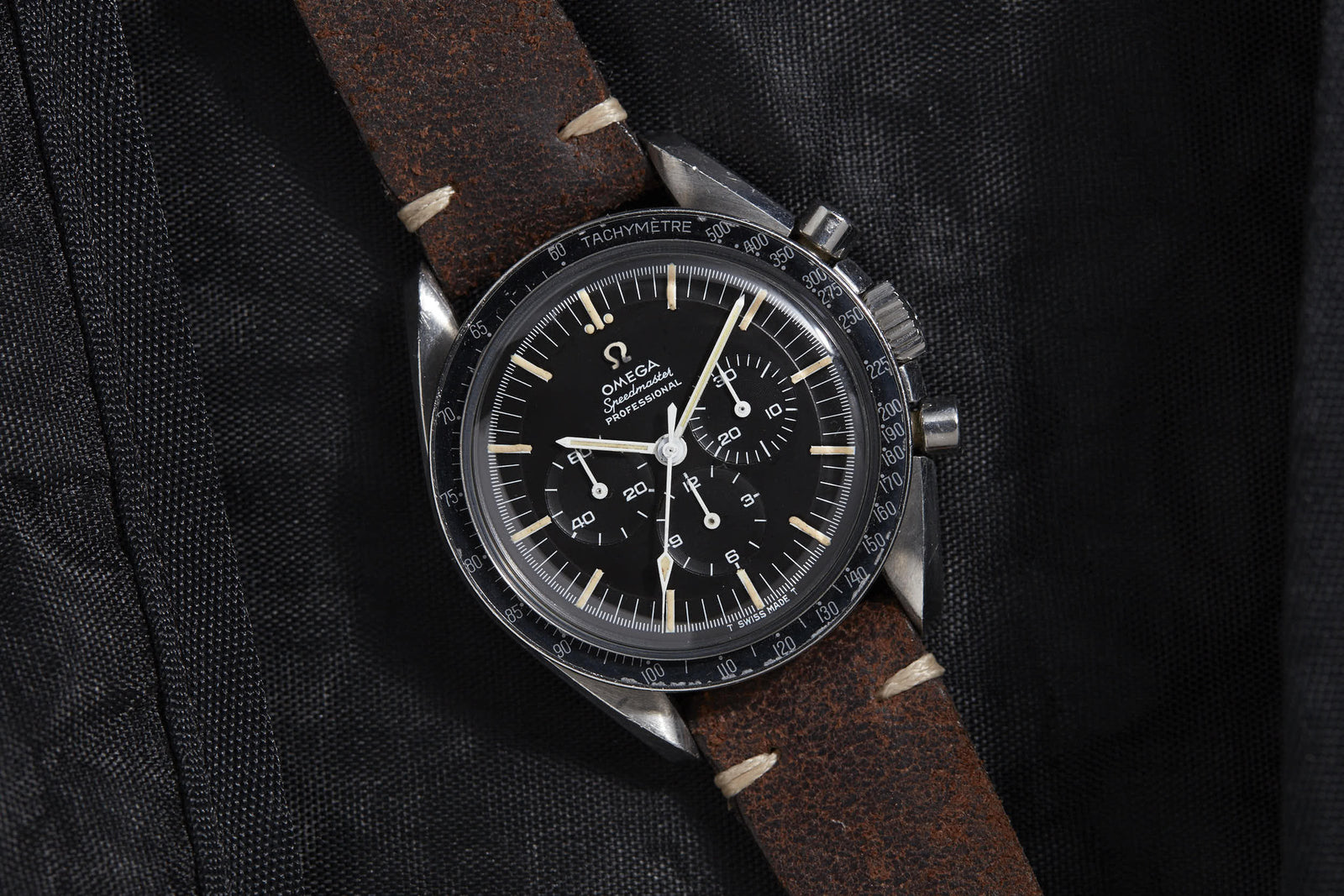 A close-up of the Omega Speedmaster Professional by Omega shows its black dial with white markers and three subdials, paired with a brown leather strap, set on textured black fabric—an iconic watch tied to NASA’s Apollo missions.